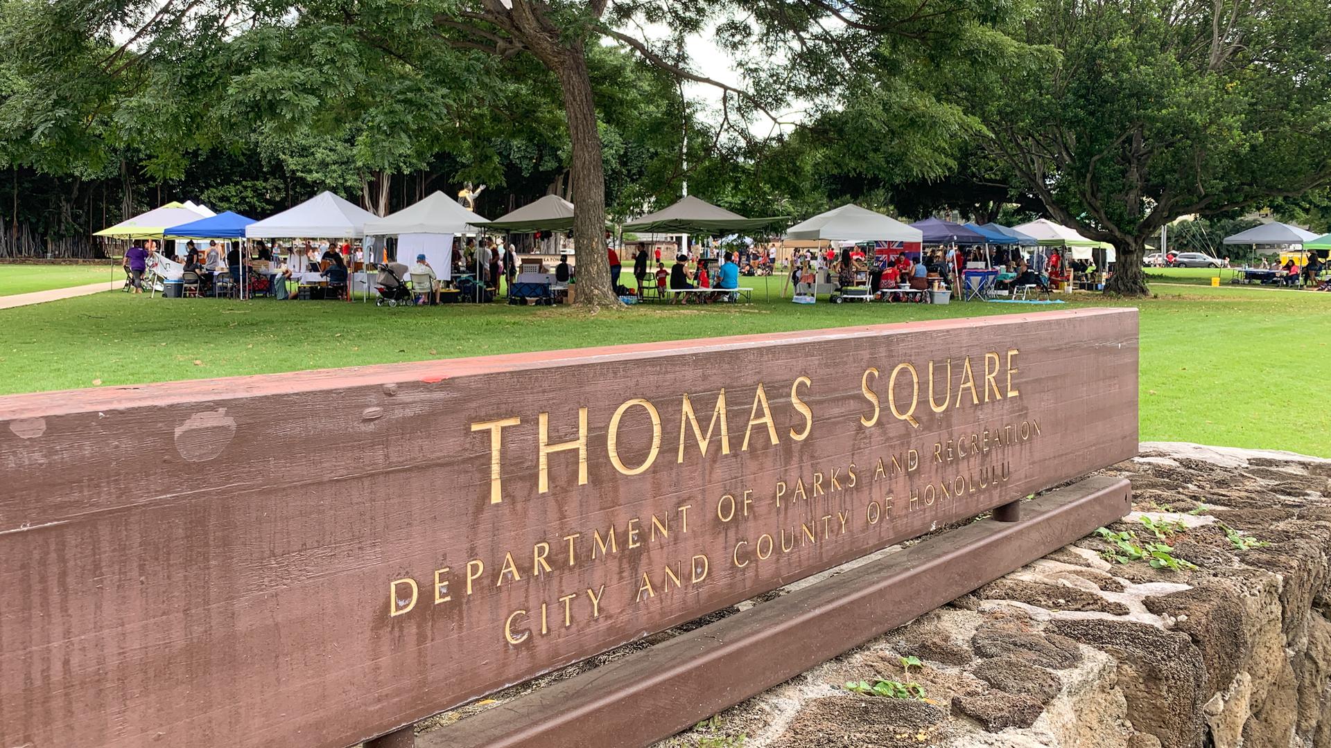 Wooden sign in foreground, outdoor market with tents in a green park.