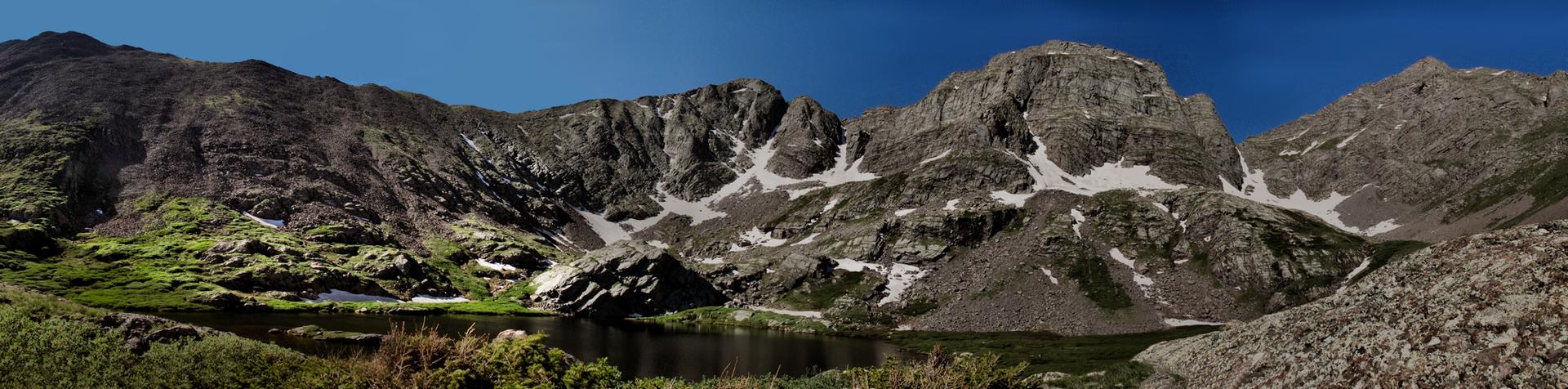 Panoramic view of rugged, snow-capped mountains and green slopes overlooking a dark lake.