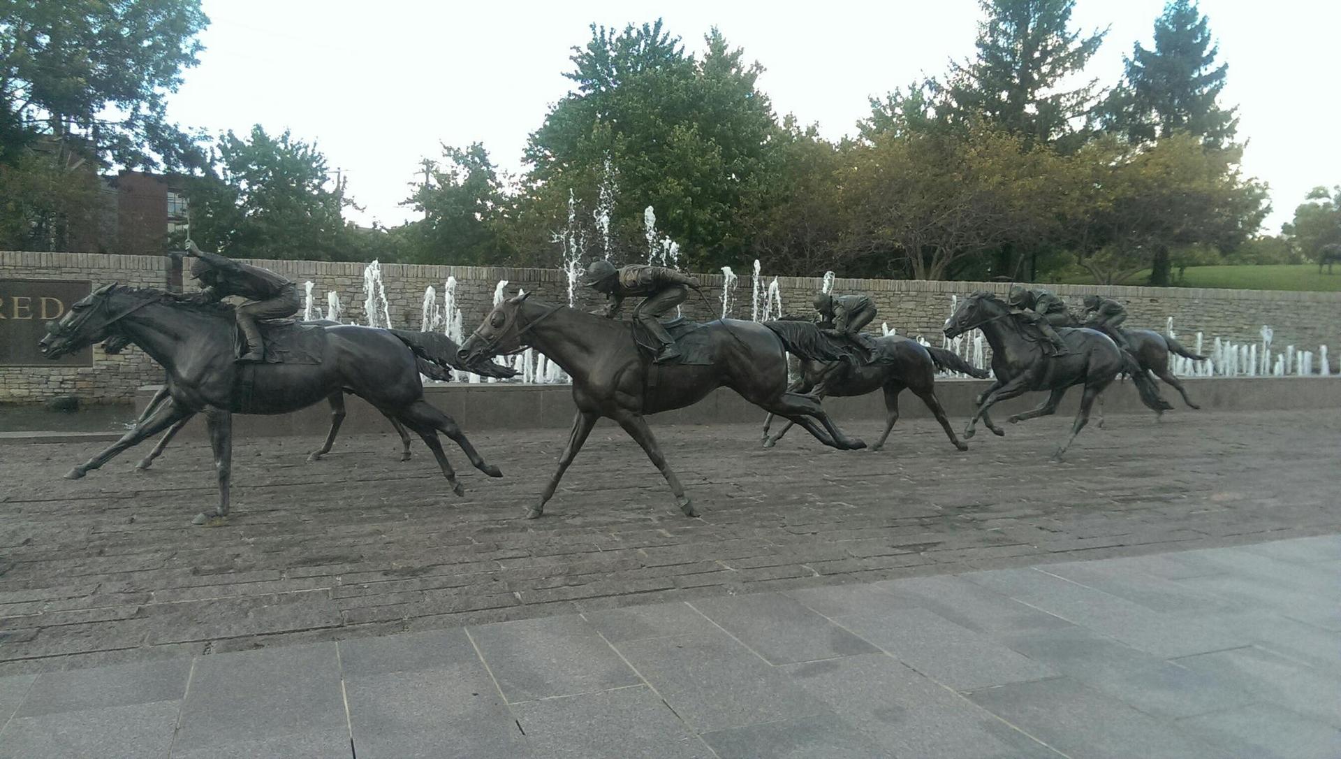 Bronze statues of jockeys racing horses with fountains behind them.