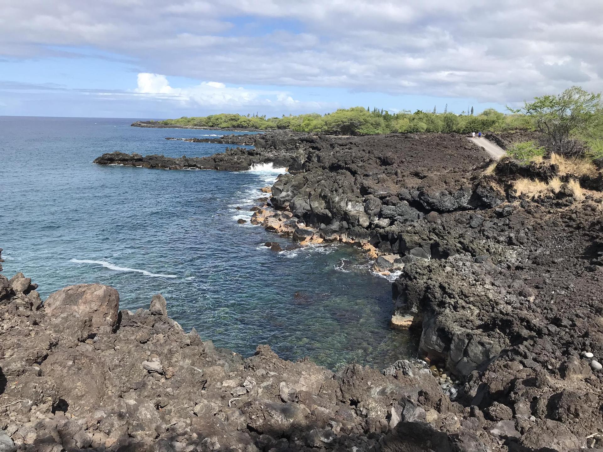 Rocky volcanic coastline with clear blue ocean under a partly cloudy sky.