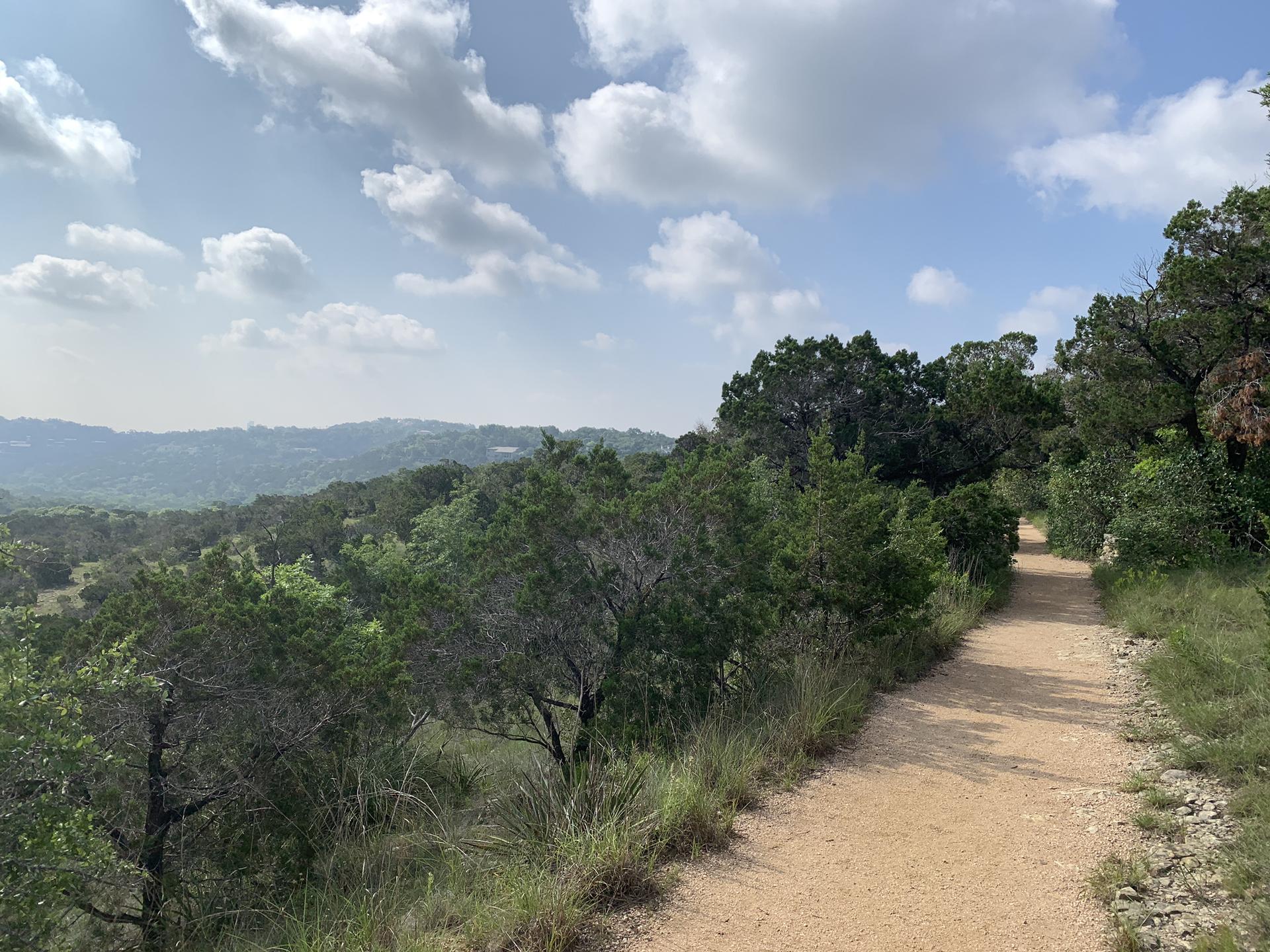 A wide dirt trail overlooks a sprawling green landscape under a bright, cloudy sky.