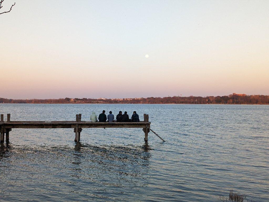 People sitting on a wooden pier over water at sunset with a visible moon.