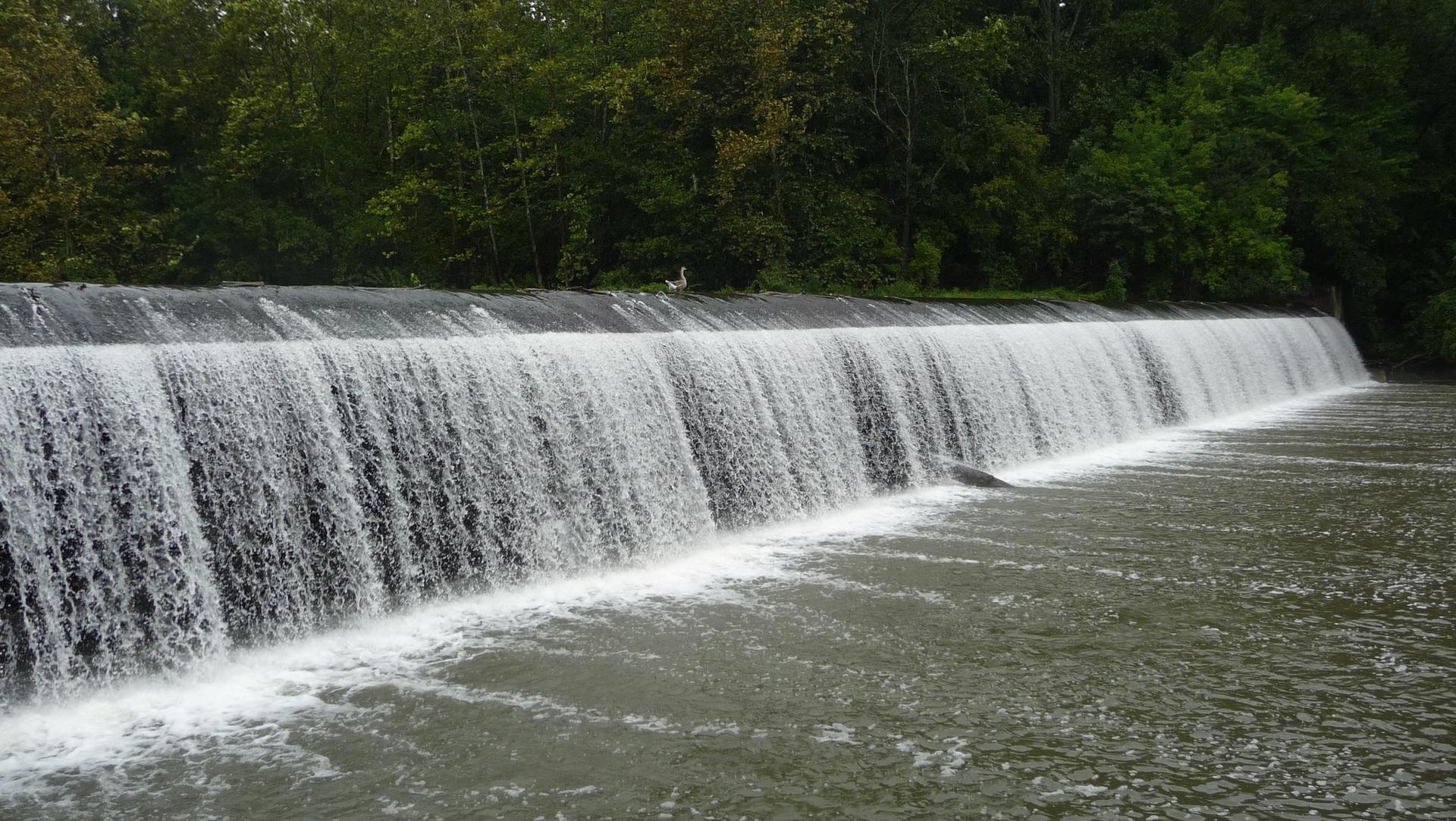 A wide waterfall cascades over a dam with green trees in the background.