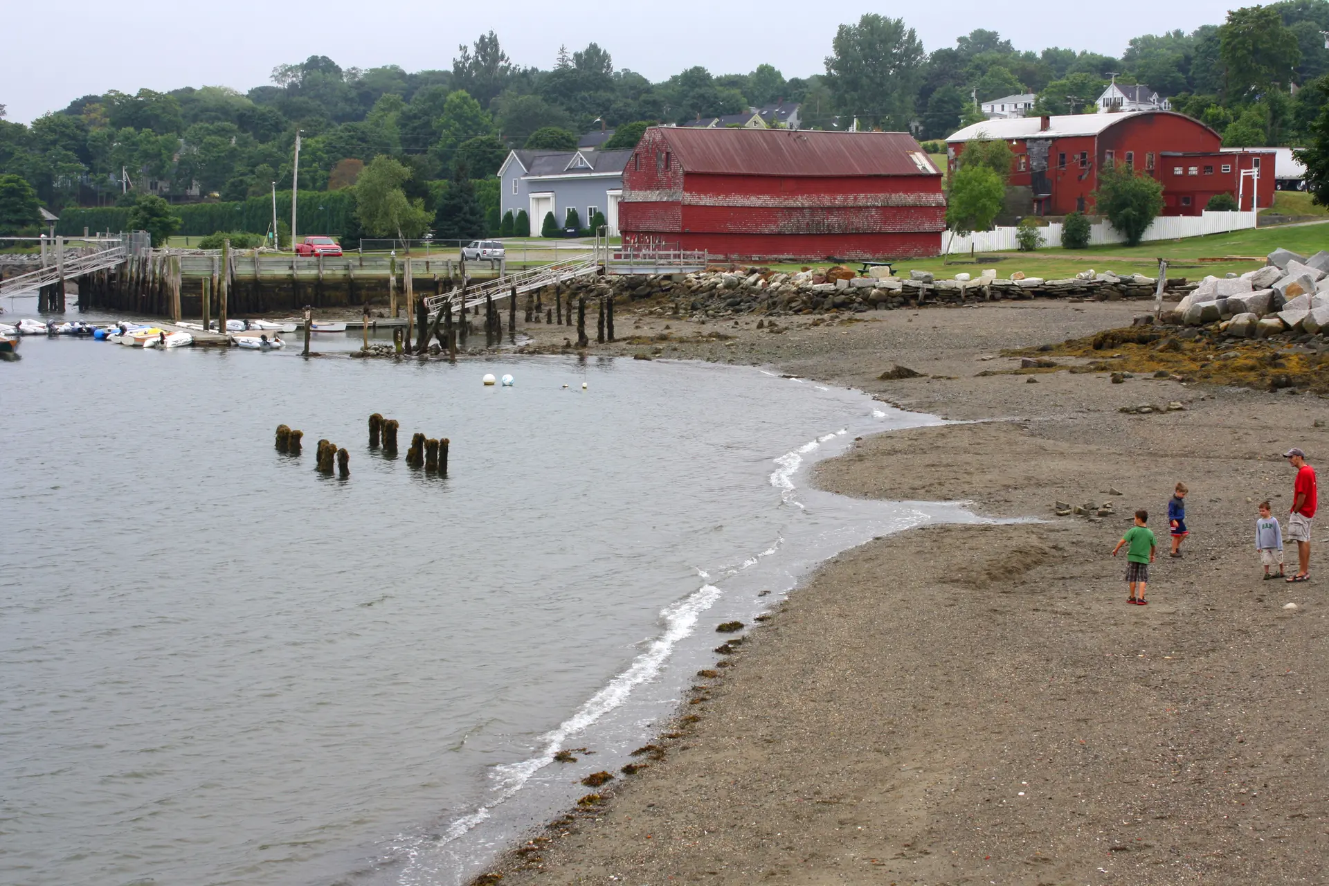 People on a sandy beach with a pier and red buildings.
