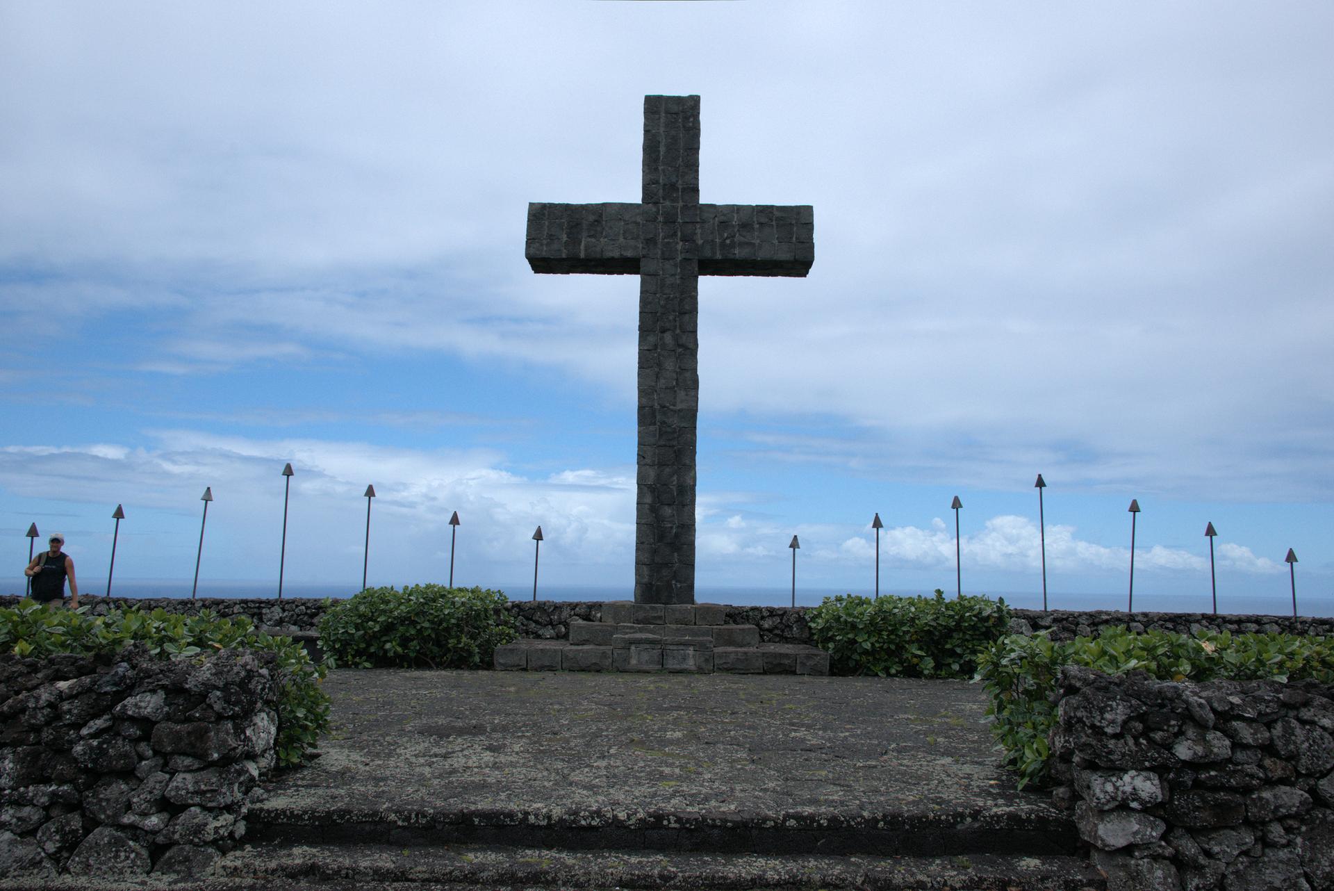 Large stone cross on a platform with spear-like posts under an overcast sky.