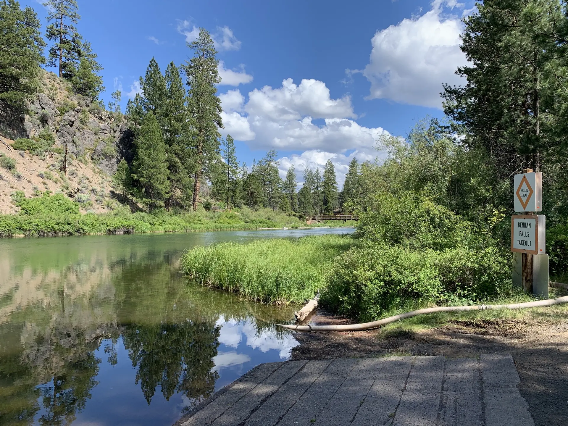 Sunny river scene with a boat launch, rocky bank, and pine trees.