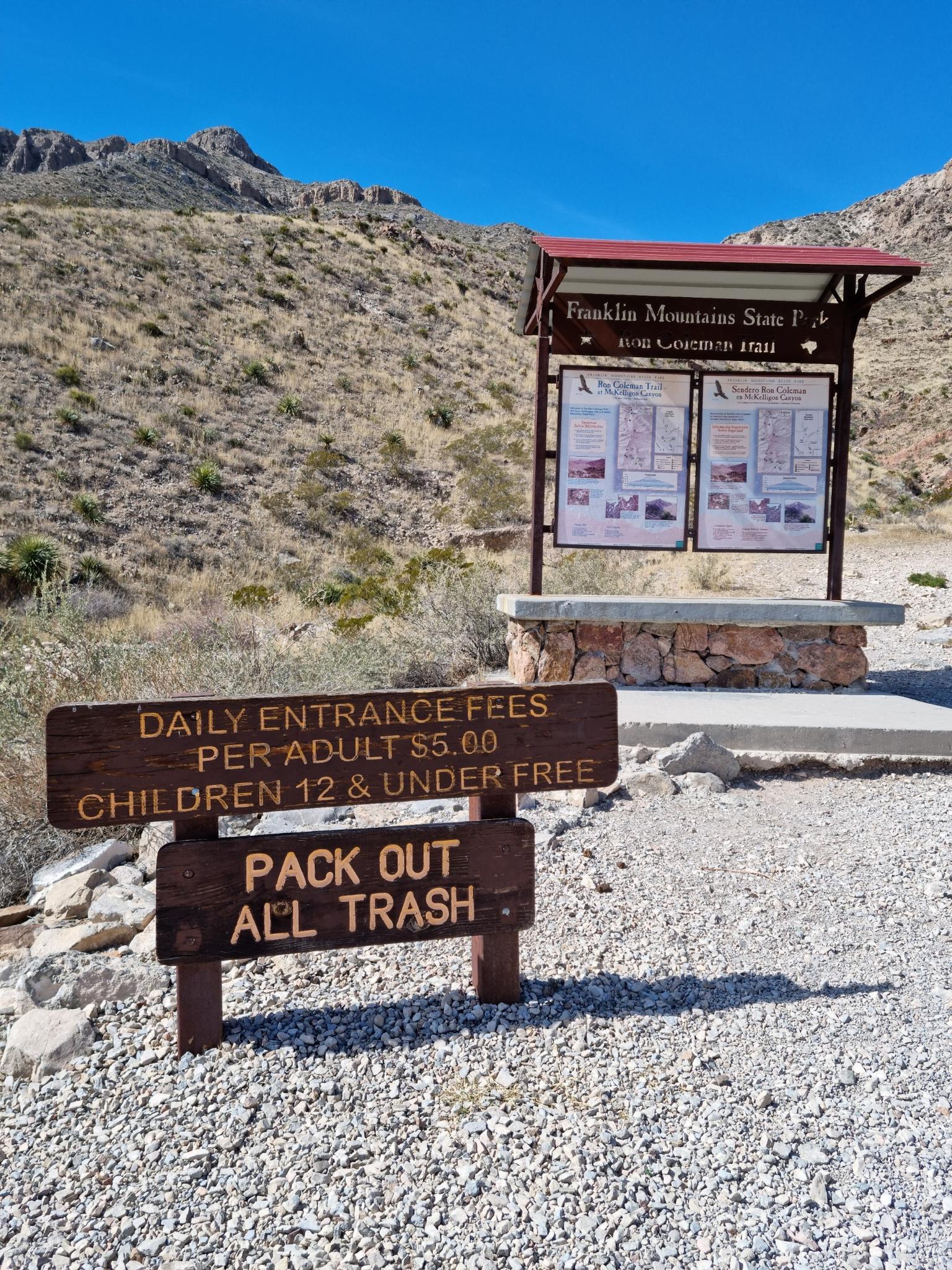 Wooden signs and an information kiosk in a sunny, arid mountain landscape.