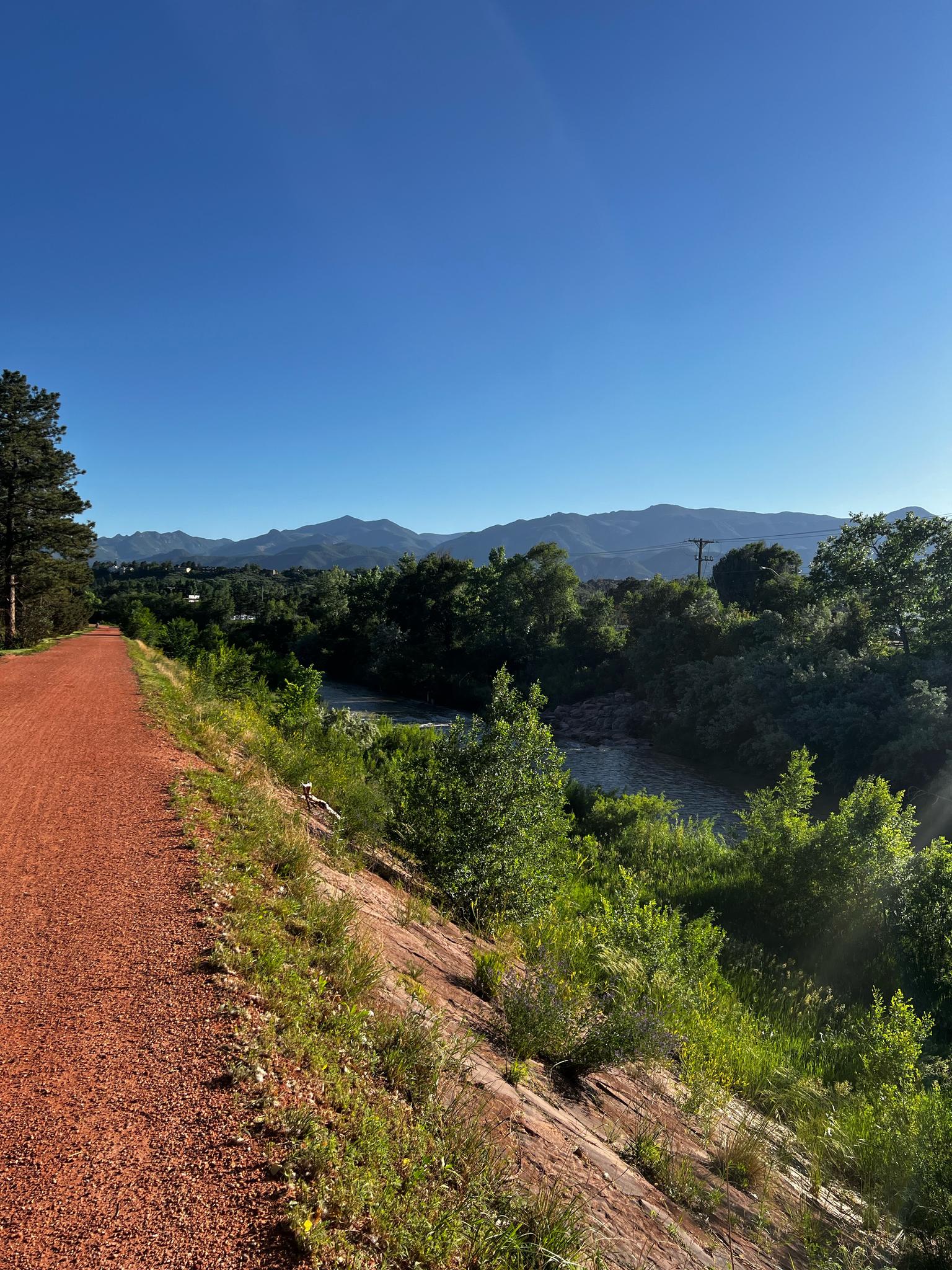 Red path beside a river and green trees, with distant mountains under clear sky.