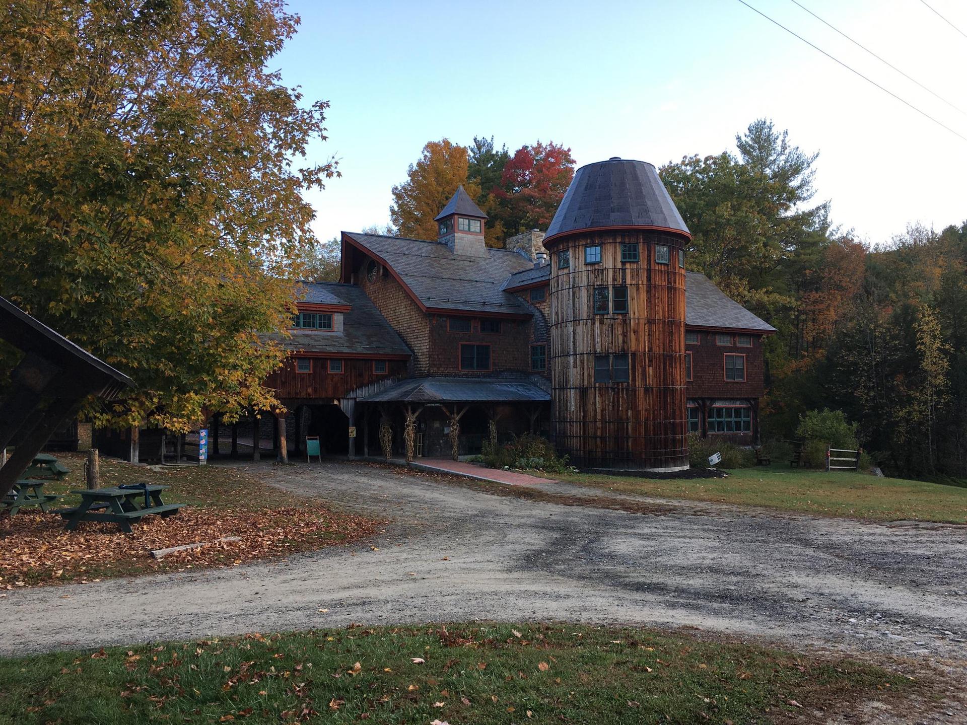 Large wooden building with a silo, surrounded by autumn trees and a gravel driveway.