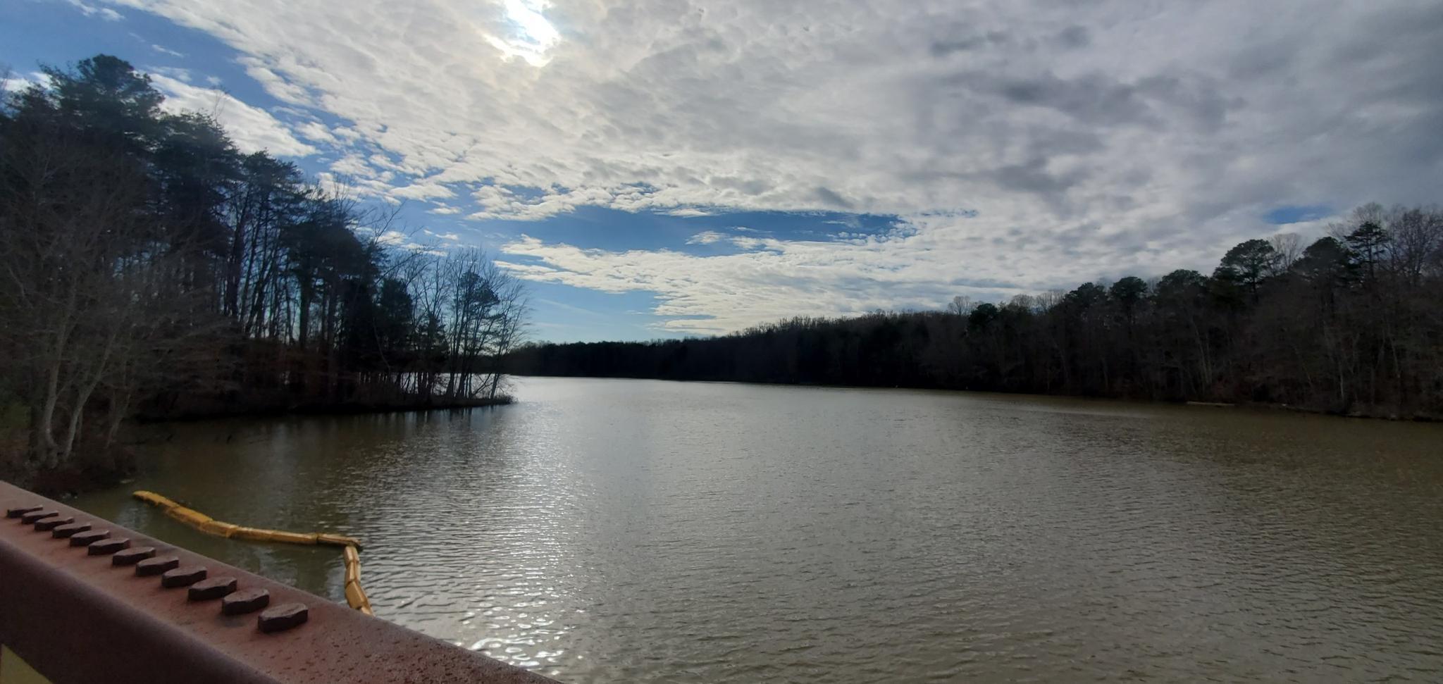 Lake with forested banks under a partly cloudy sky, rusty railing foreground.