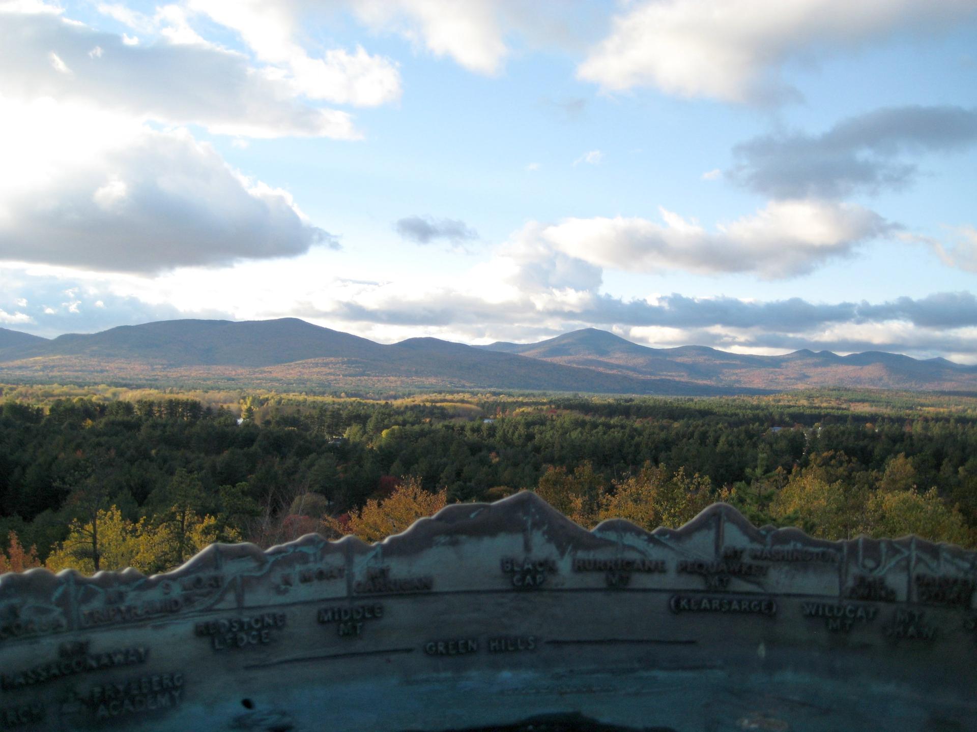 Panoramic view of mountains, autumn forest, and an interpretive plaque under a cloudy sky.