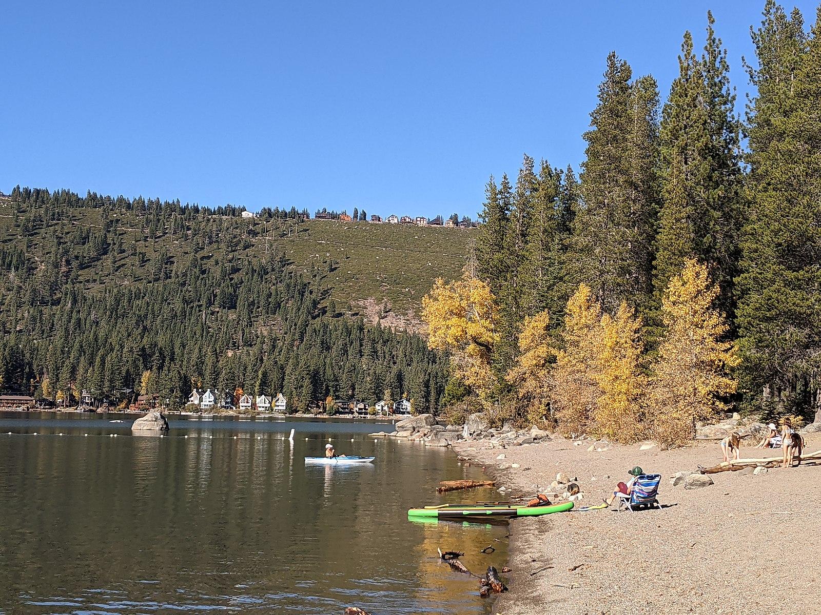 Sunny lake scene with paddleboarders, people on a sandy beach, and autumn trees.