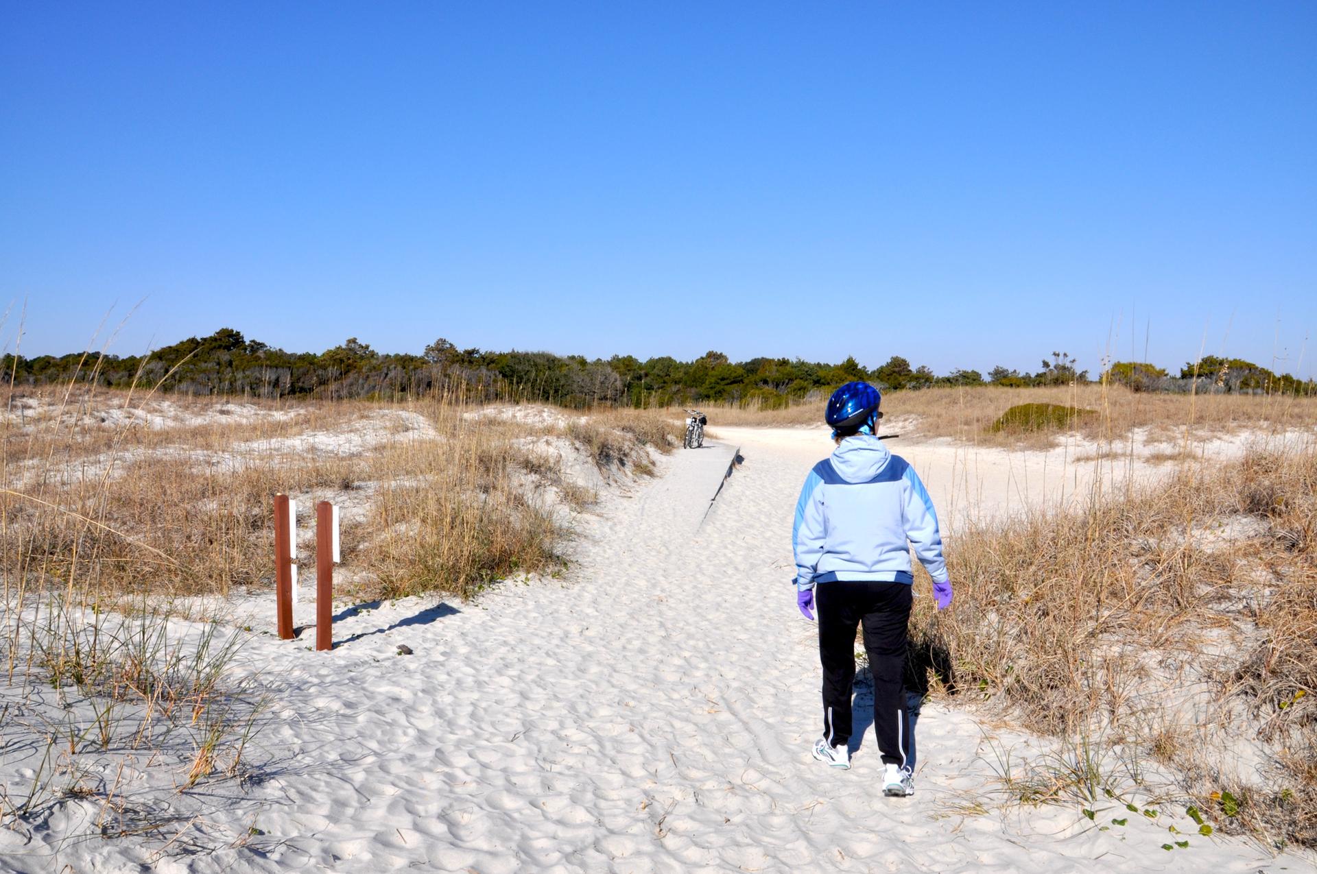 A person walks on a sandy path through dunes under a clear blue sky.