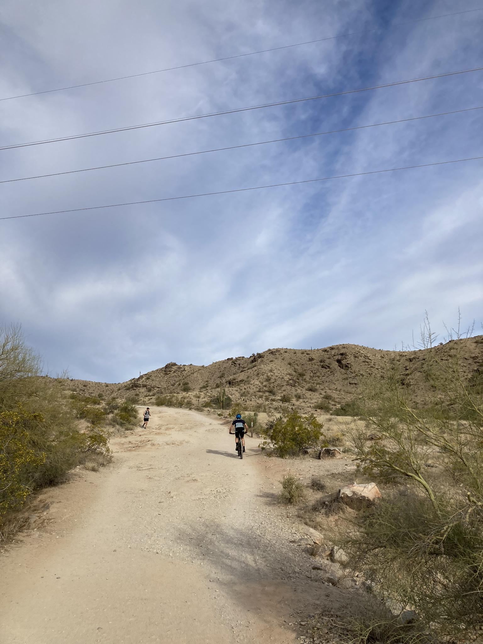 Cyclist and walker on a desert trail under a bright, cloudy sky.