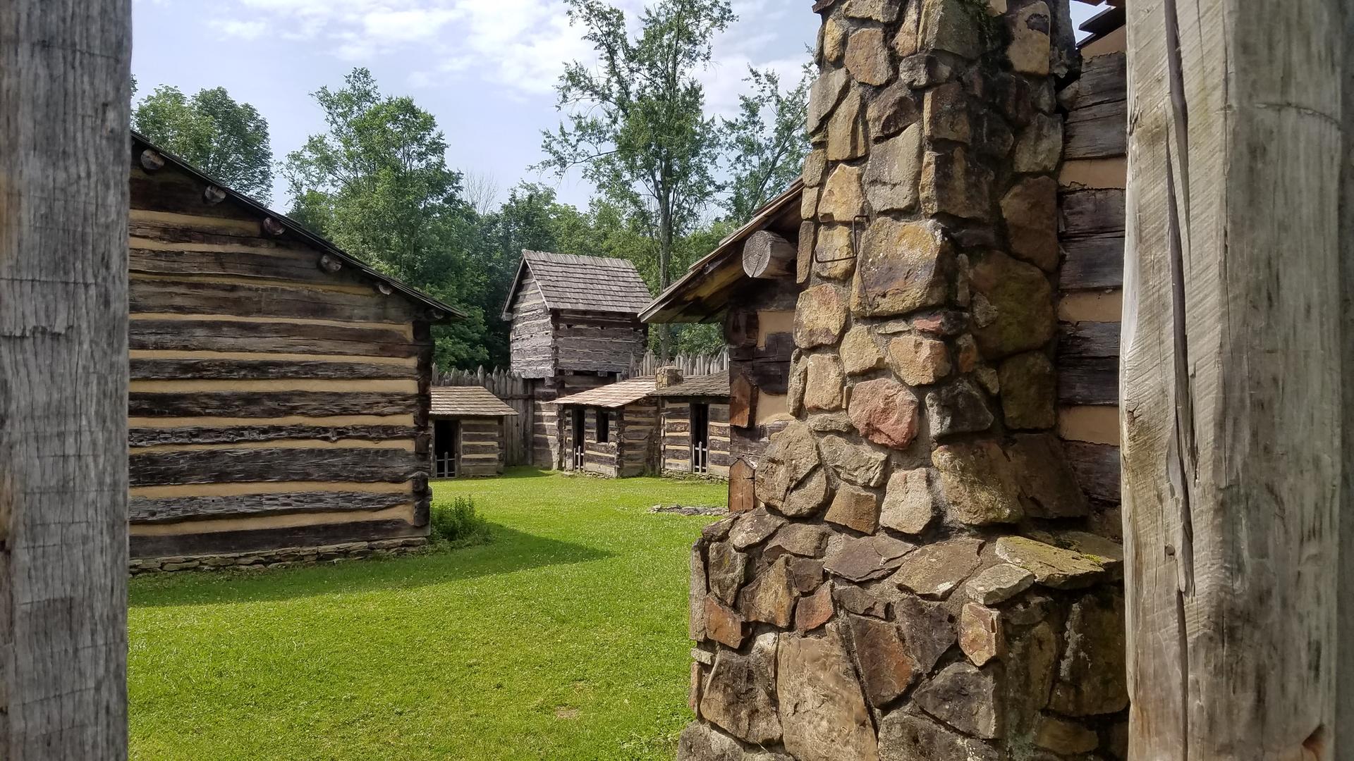 Historic log cabins with a stone chimney in a grassy clearing on a cloudy day.