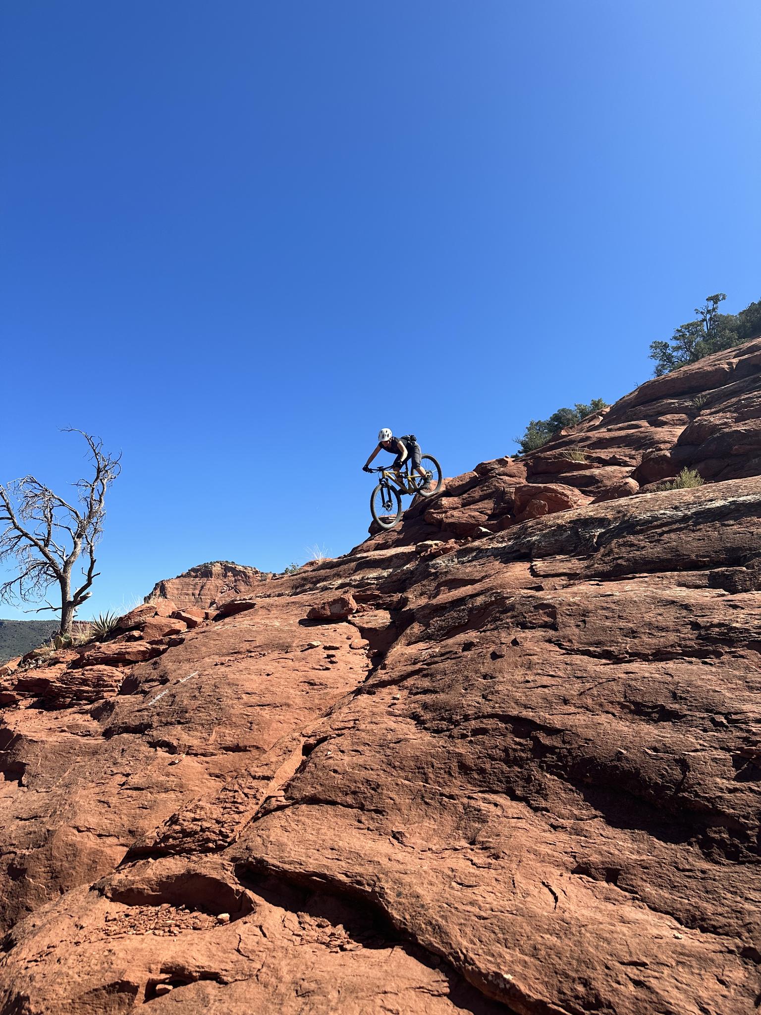 Mountain biker descends a steep, rocky red slope under a clear blue sky.