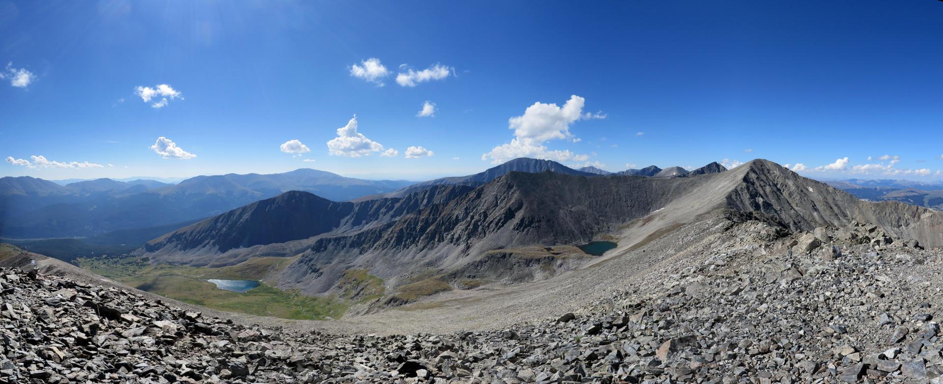 Panoramic view of rocky mountain peaks and two alpine lakes under a bright sky.