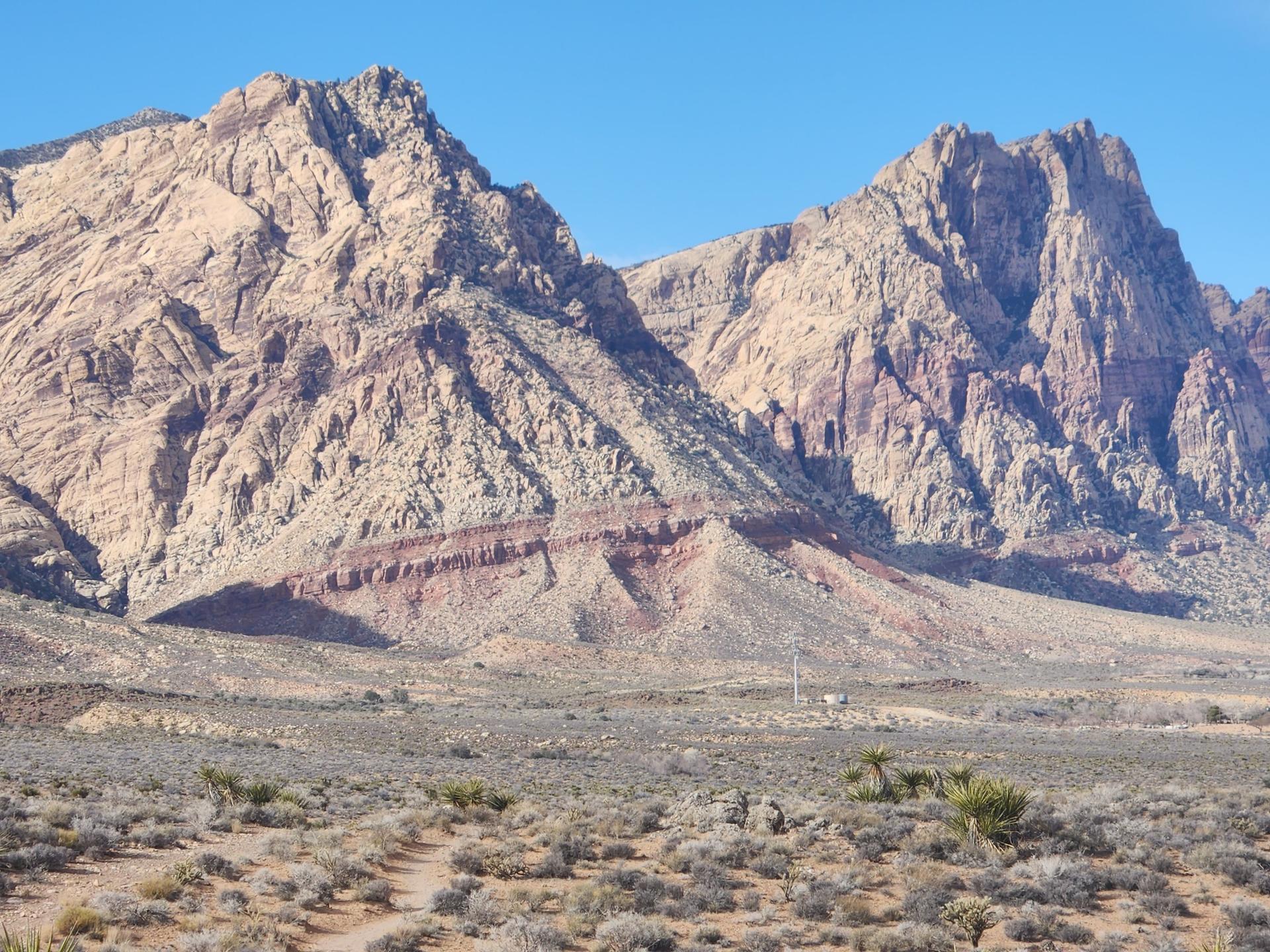 Massive, layered desert mountains tower over a scrub-filled landscape on a sunny day.