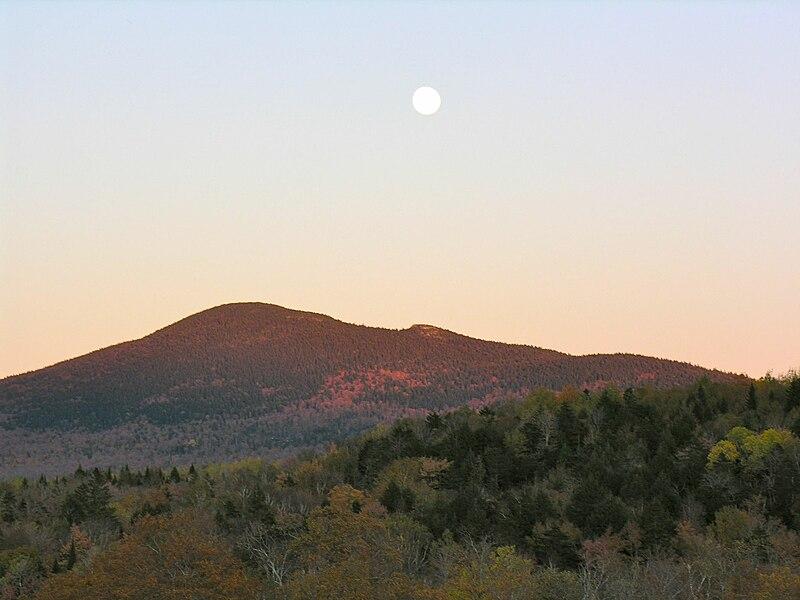 A full moon hangs over forested mountains with autumn colors at dusk.