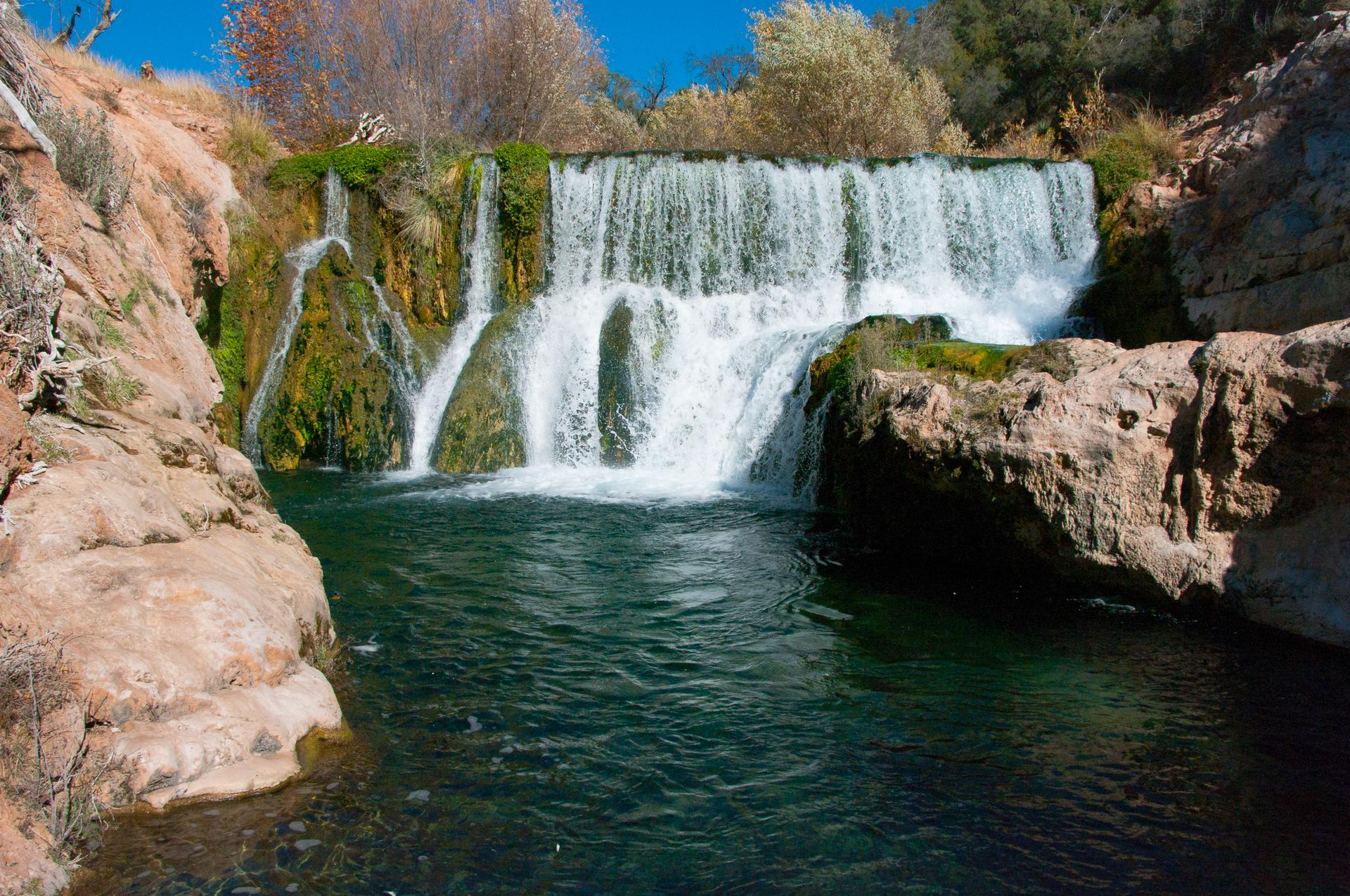 A multi-tiered waterfall cascades into a clear, dark blue pool surrounded by rocks.