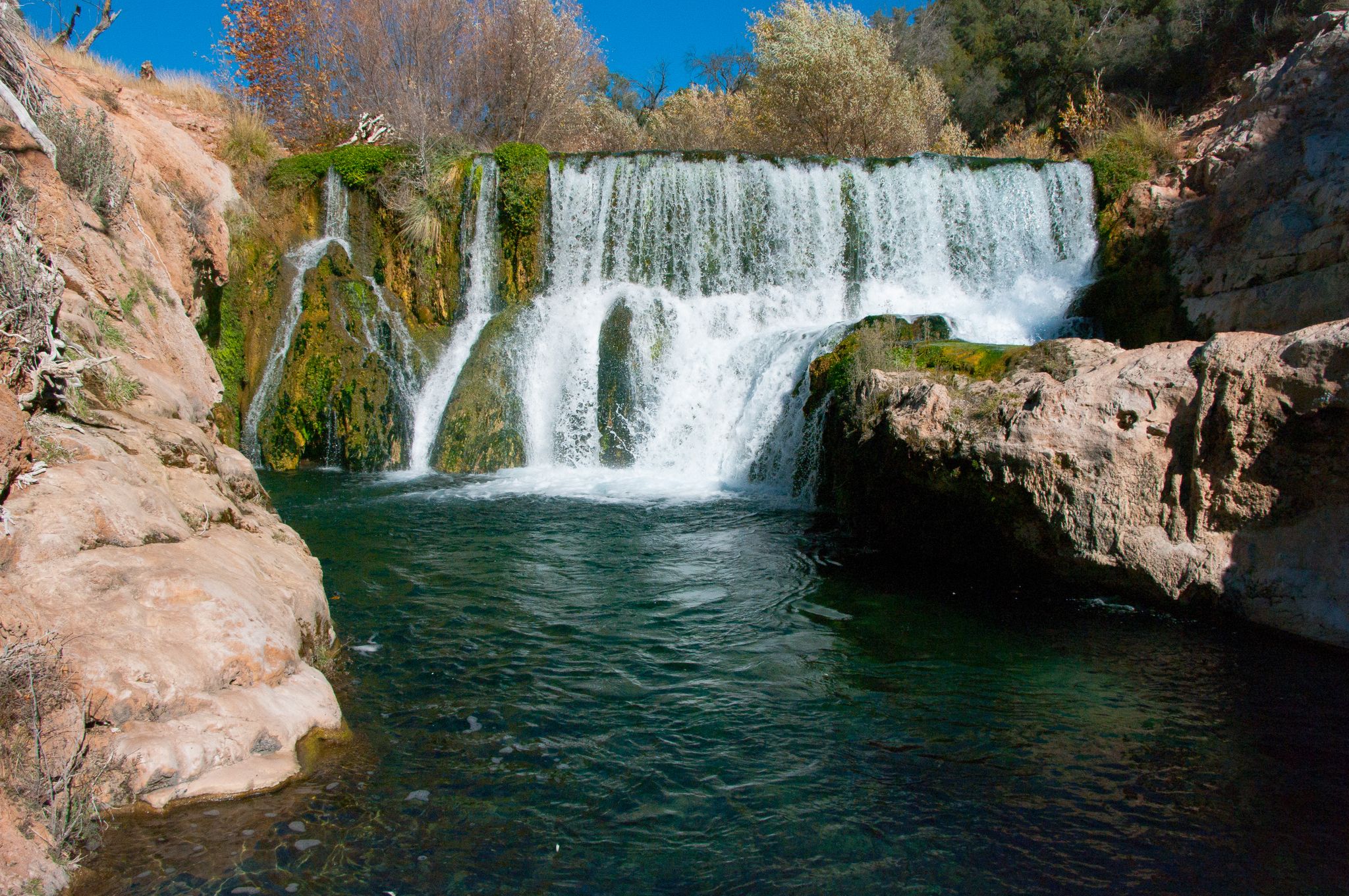 A multi-tiered waterfall cascades into a clear, dark blue pool surrounded by rocks. - Bob Bear Trail