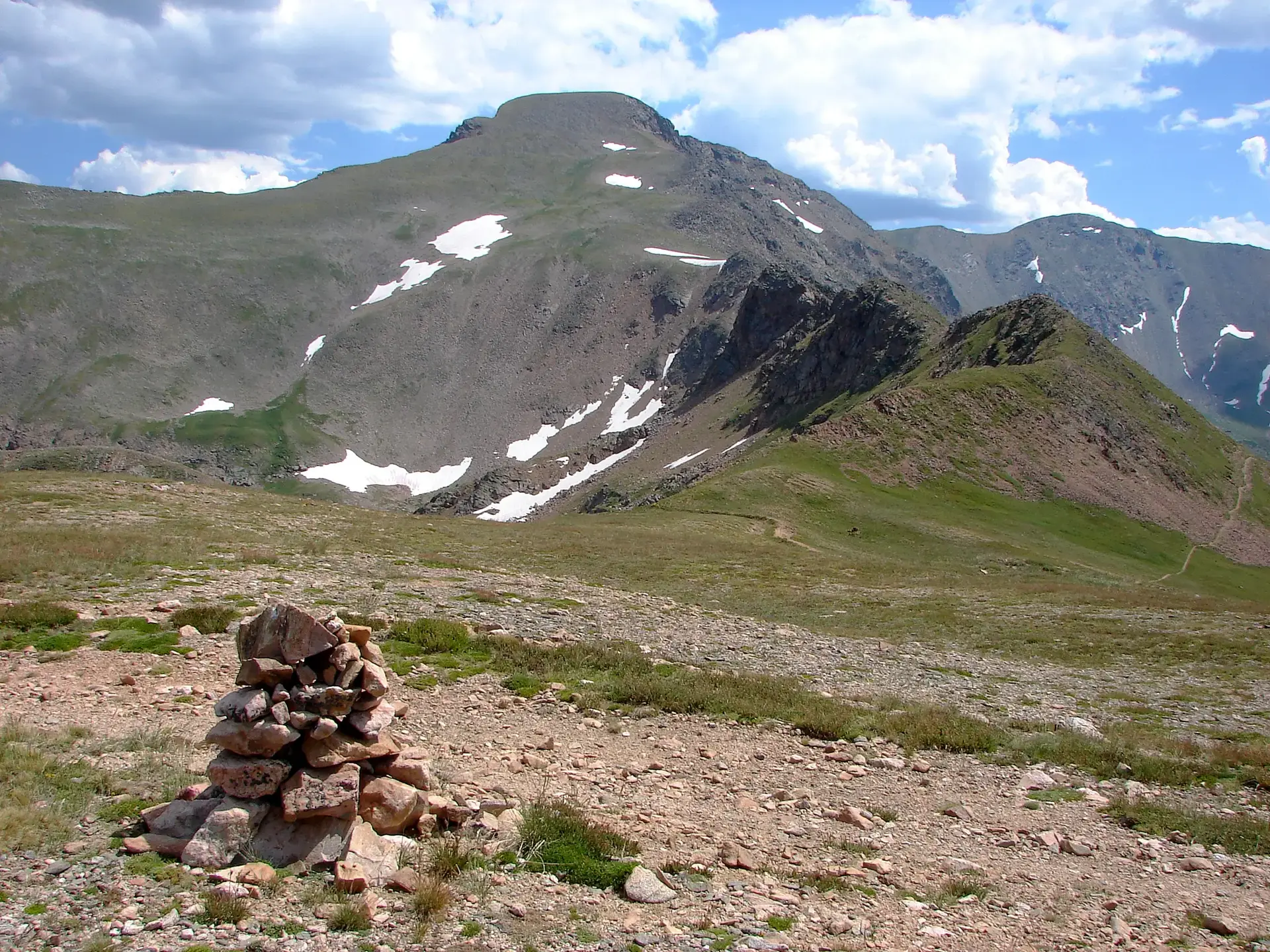 A rock cairn on a rocky path with snow-dusted mountains under a bright, cloudy sky.