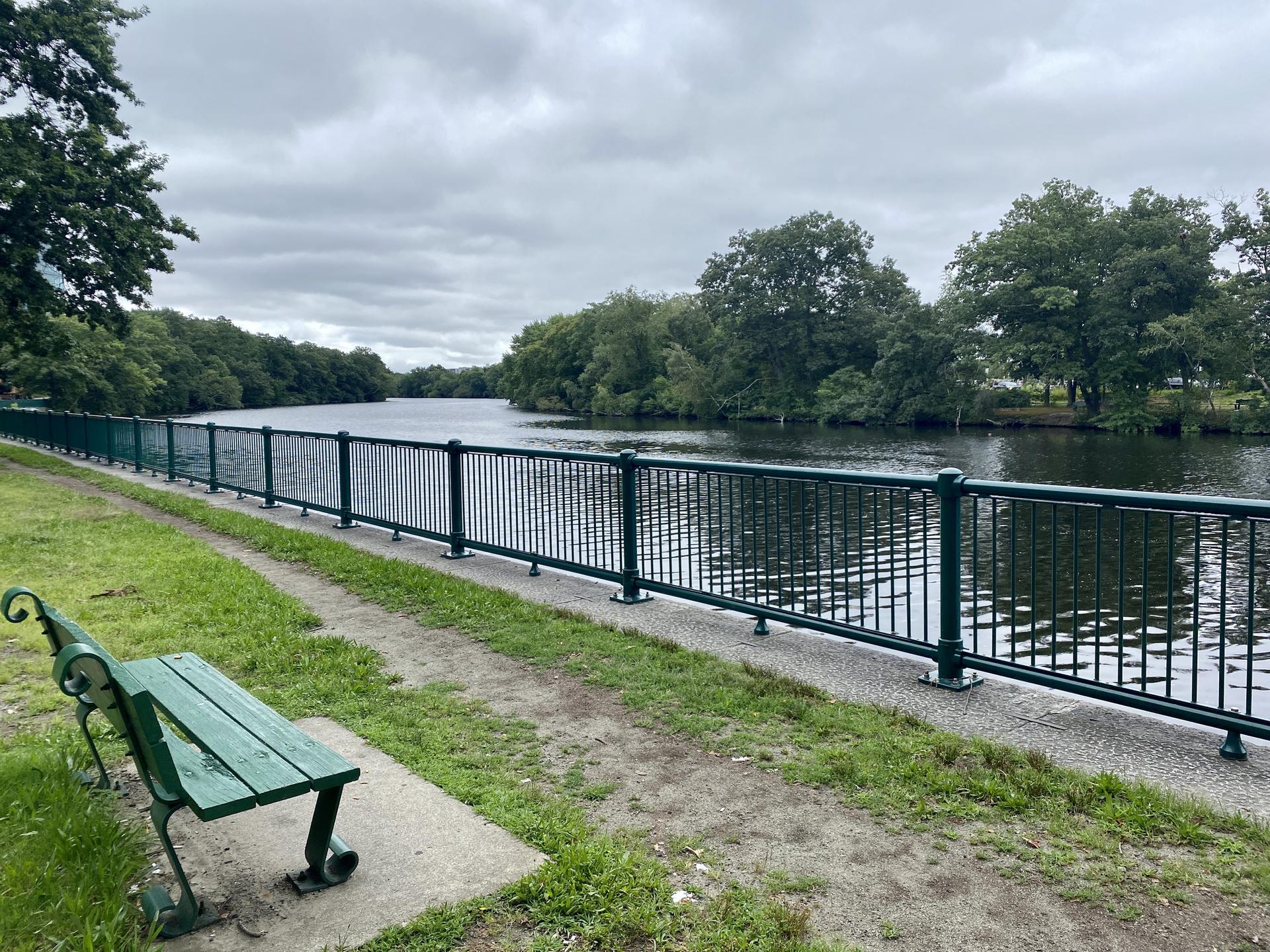 Green park bench beside a river with a fence on an overcast day.