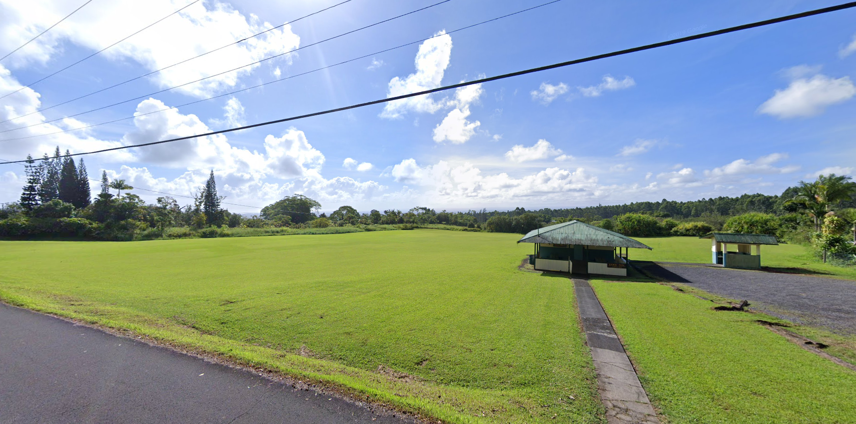 Sunny green field with two small buildings and distant trees under blue sky.