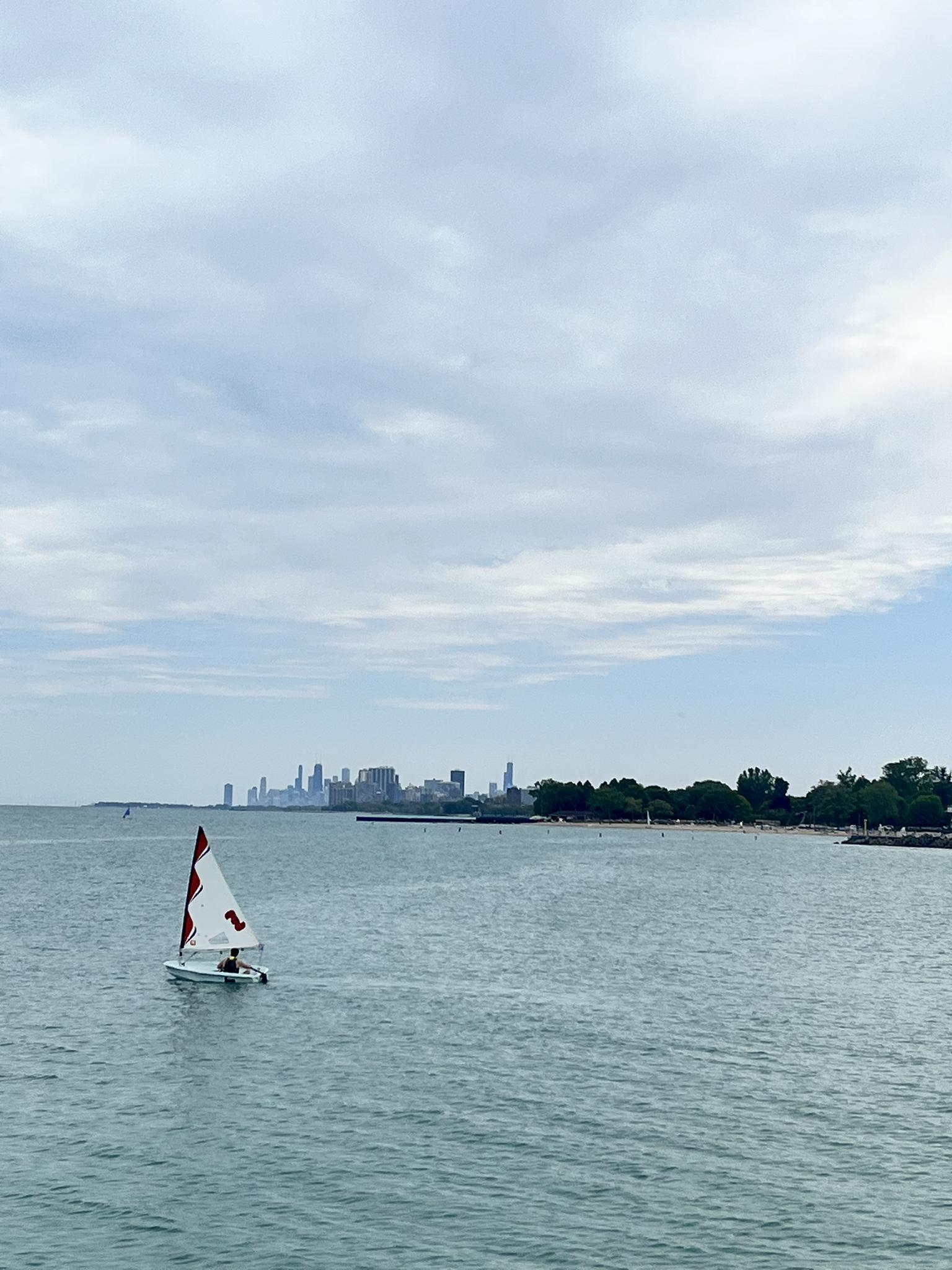 A person sails a small boat on calm water with a distant city skyline.