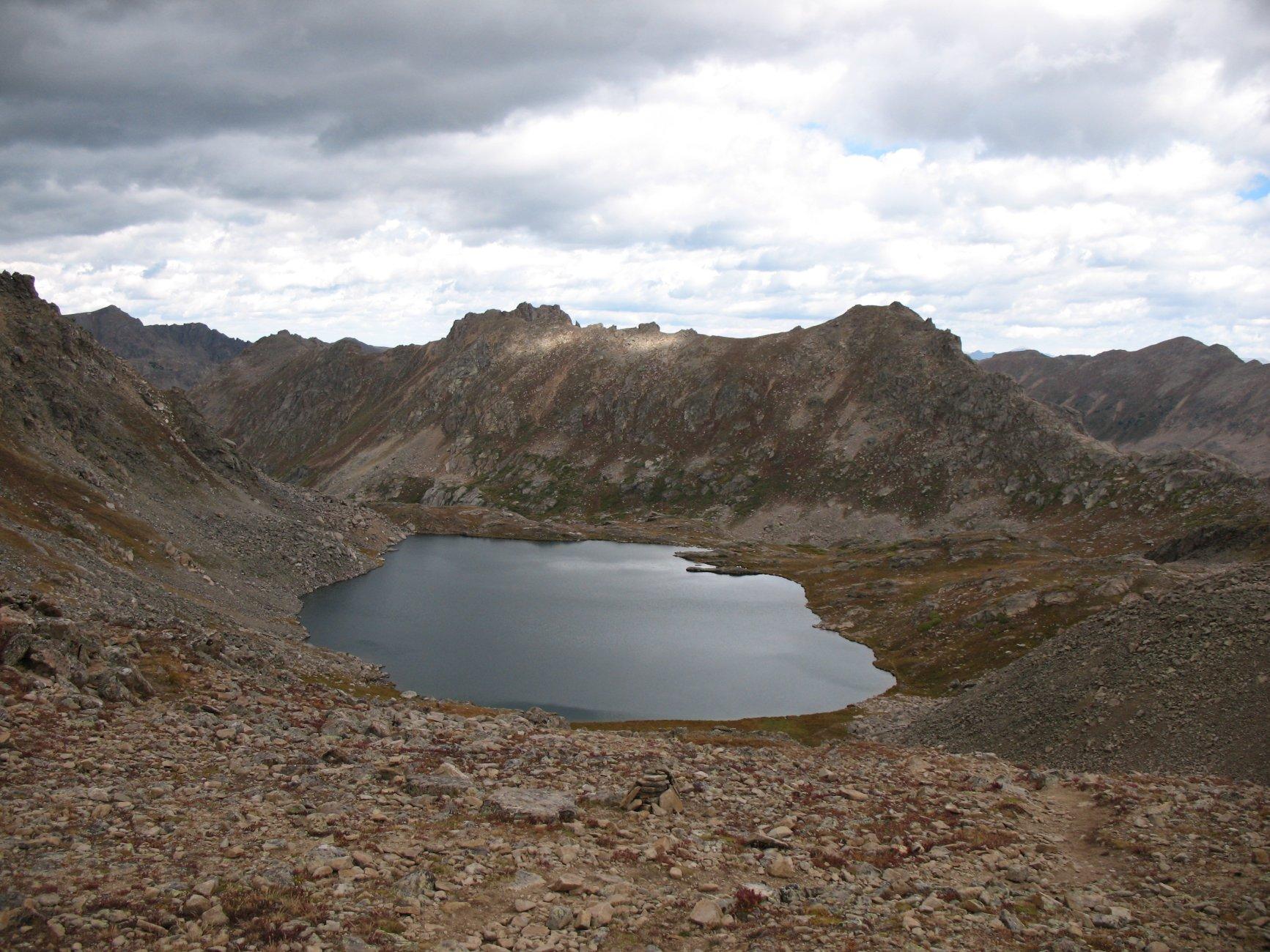 Dark mountain lake nestled in barren, rocky terrain under dramatic, cloudy skies.