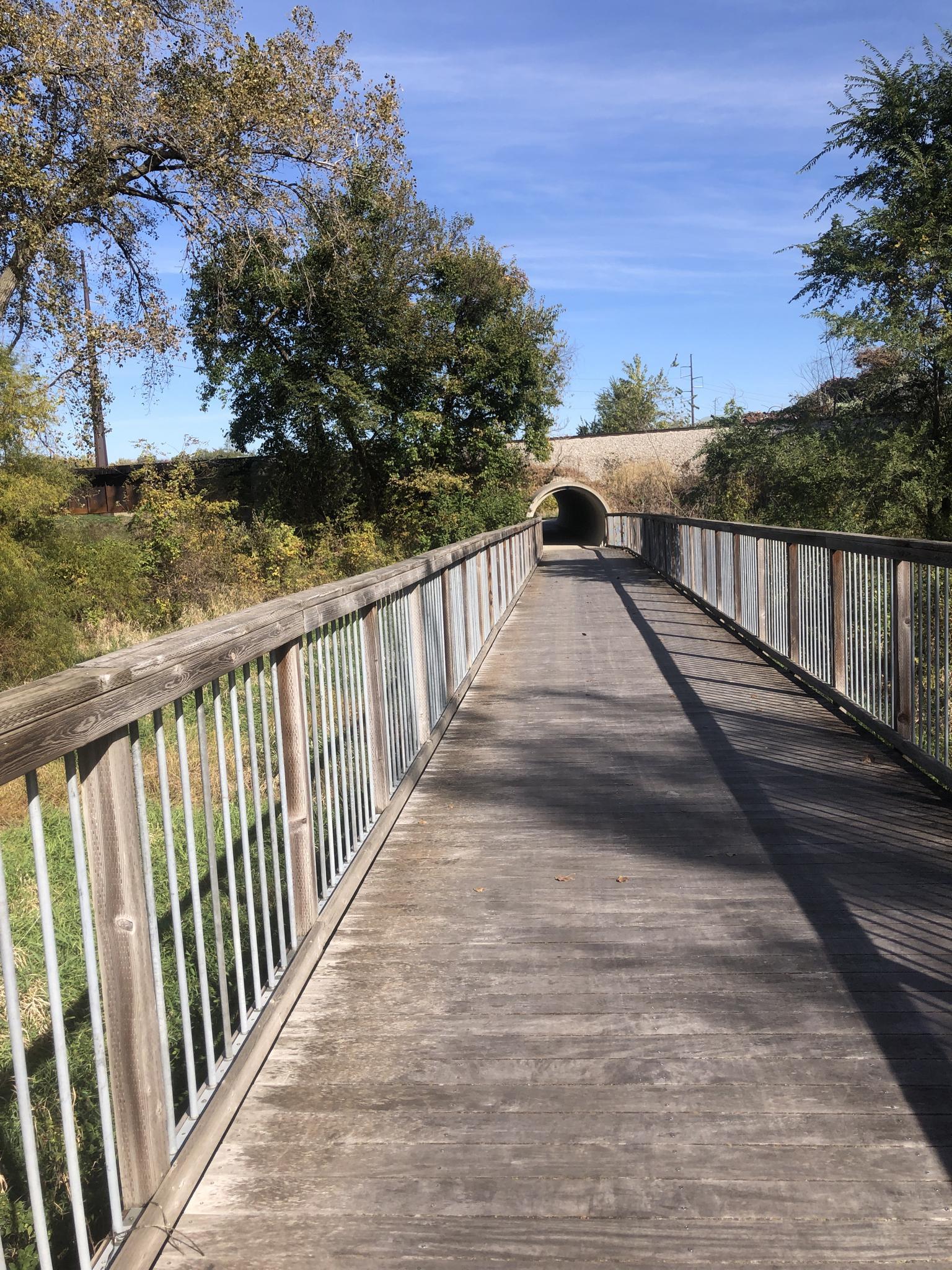 Wooden boardwalk leading to a tunnel under a bridge on a sunny day.