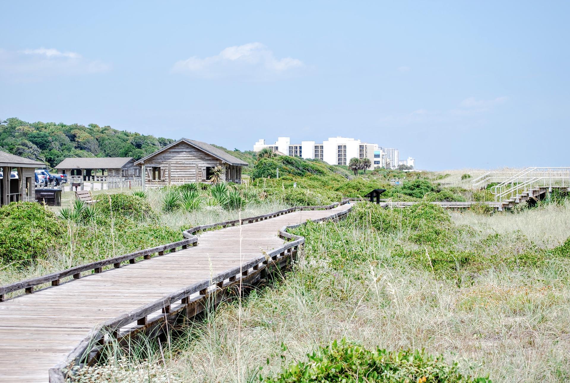 Wooden boardwalk winding through grassy dunes towards distant buildings under a bright sky.