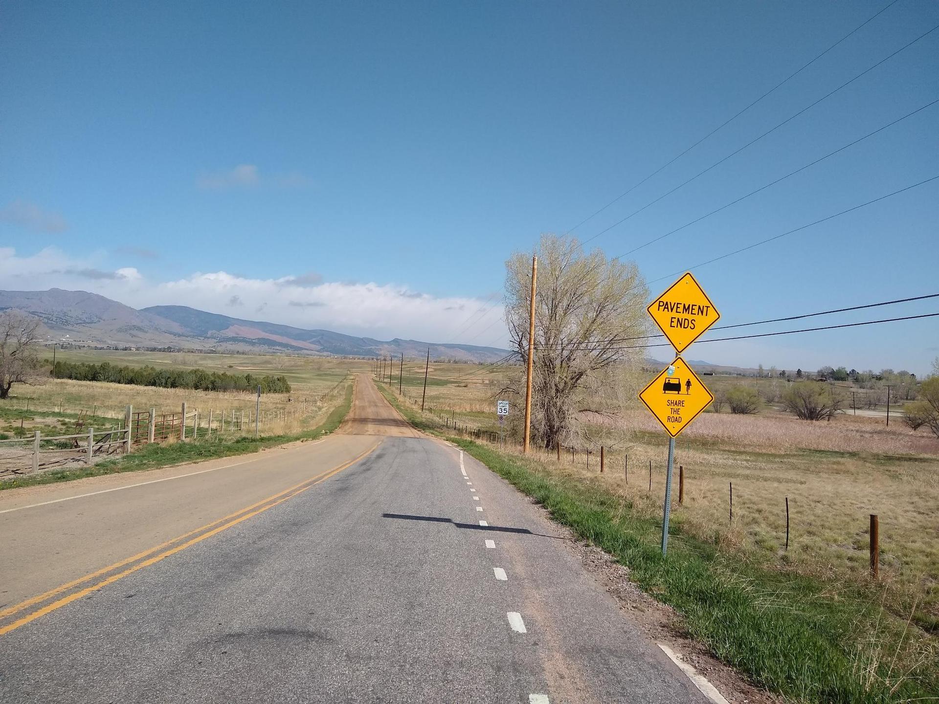 Paved road ends, becoming dirt, with warning signs under a clear sky.