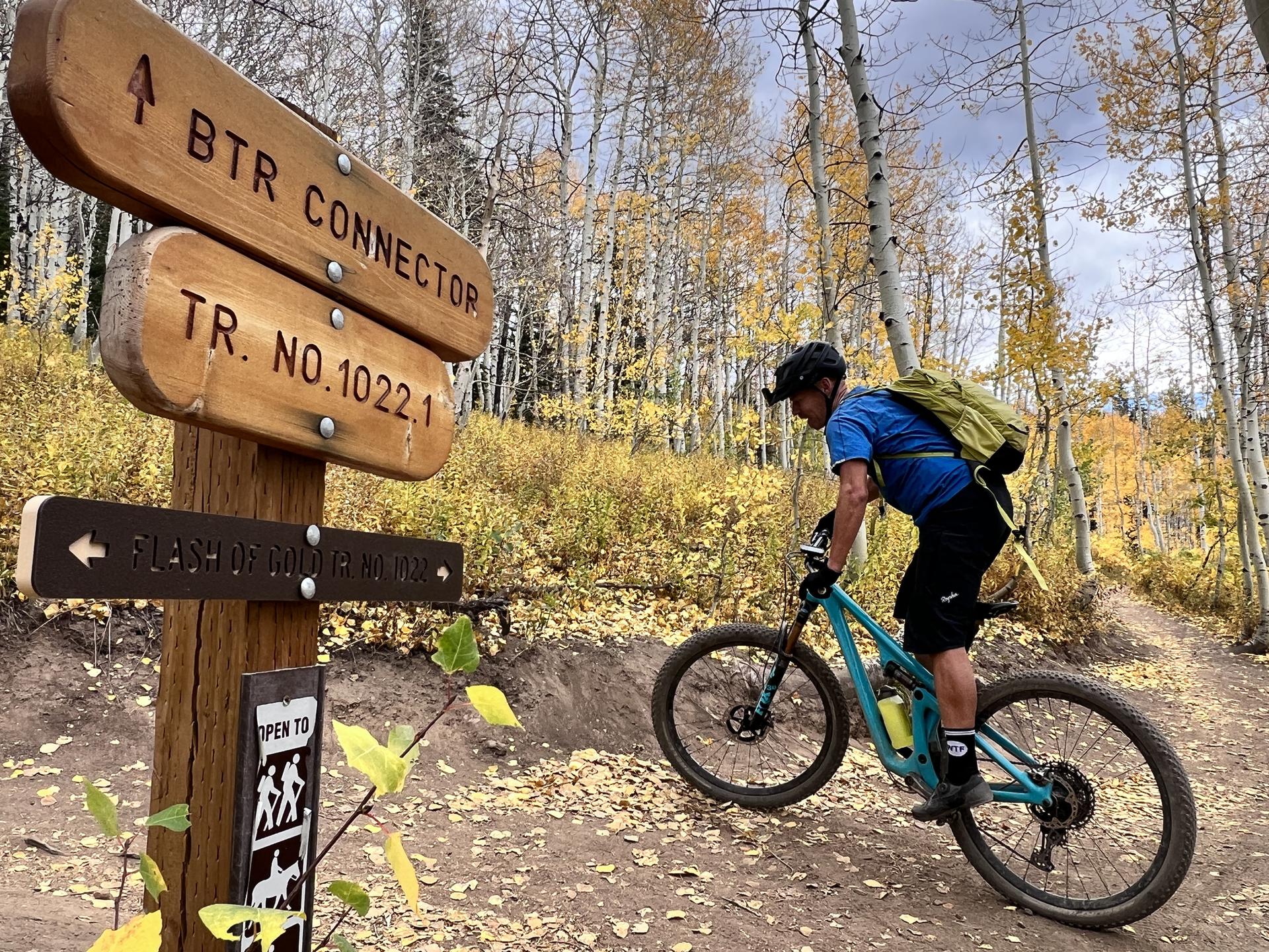 Mountain biker pauses by a wooden trail sign in a golden autumn forest.
