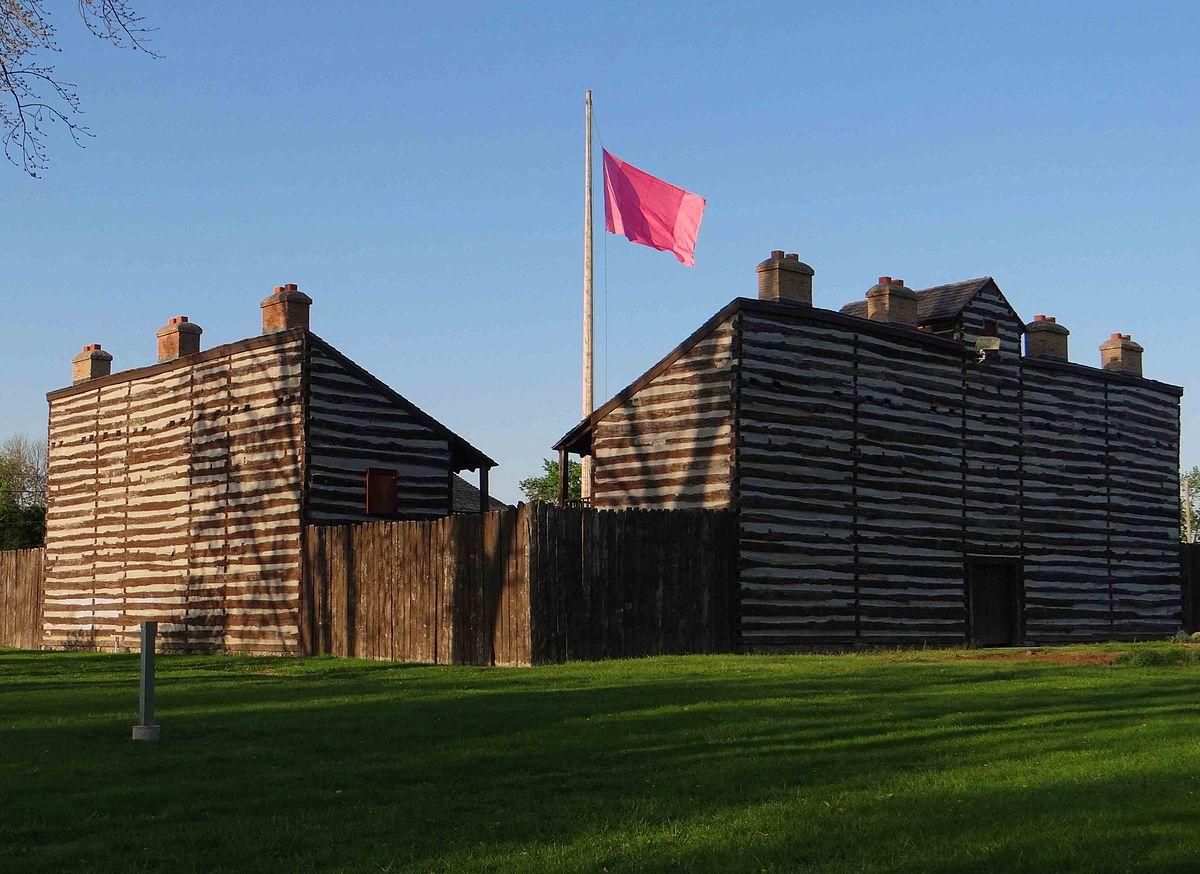Two historic log buildings with a pink flag flying on a sunny evening.