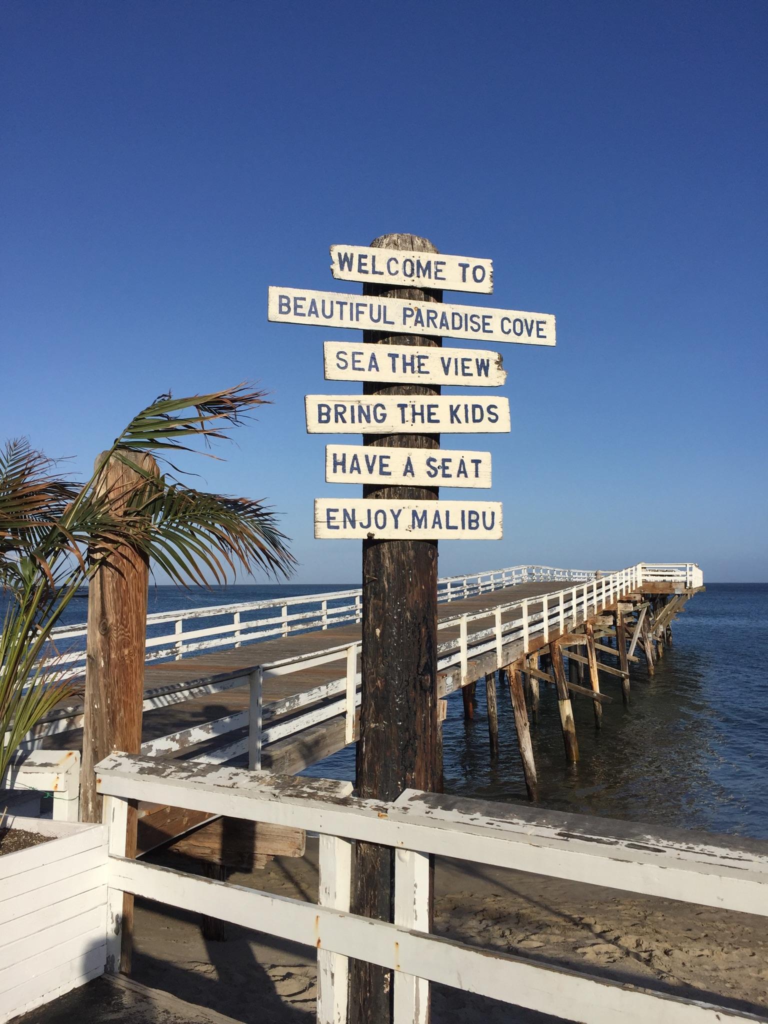 Wooden signpost with beach directions, pier extending into ocean, under a clear sky.