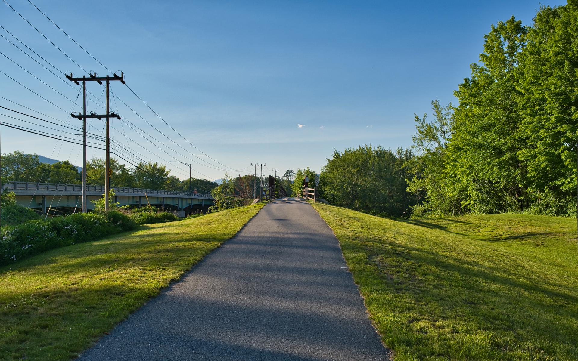 A paved path flanked by green grass and trees under a bright blue sky.