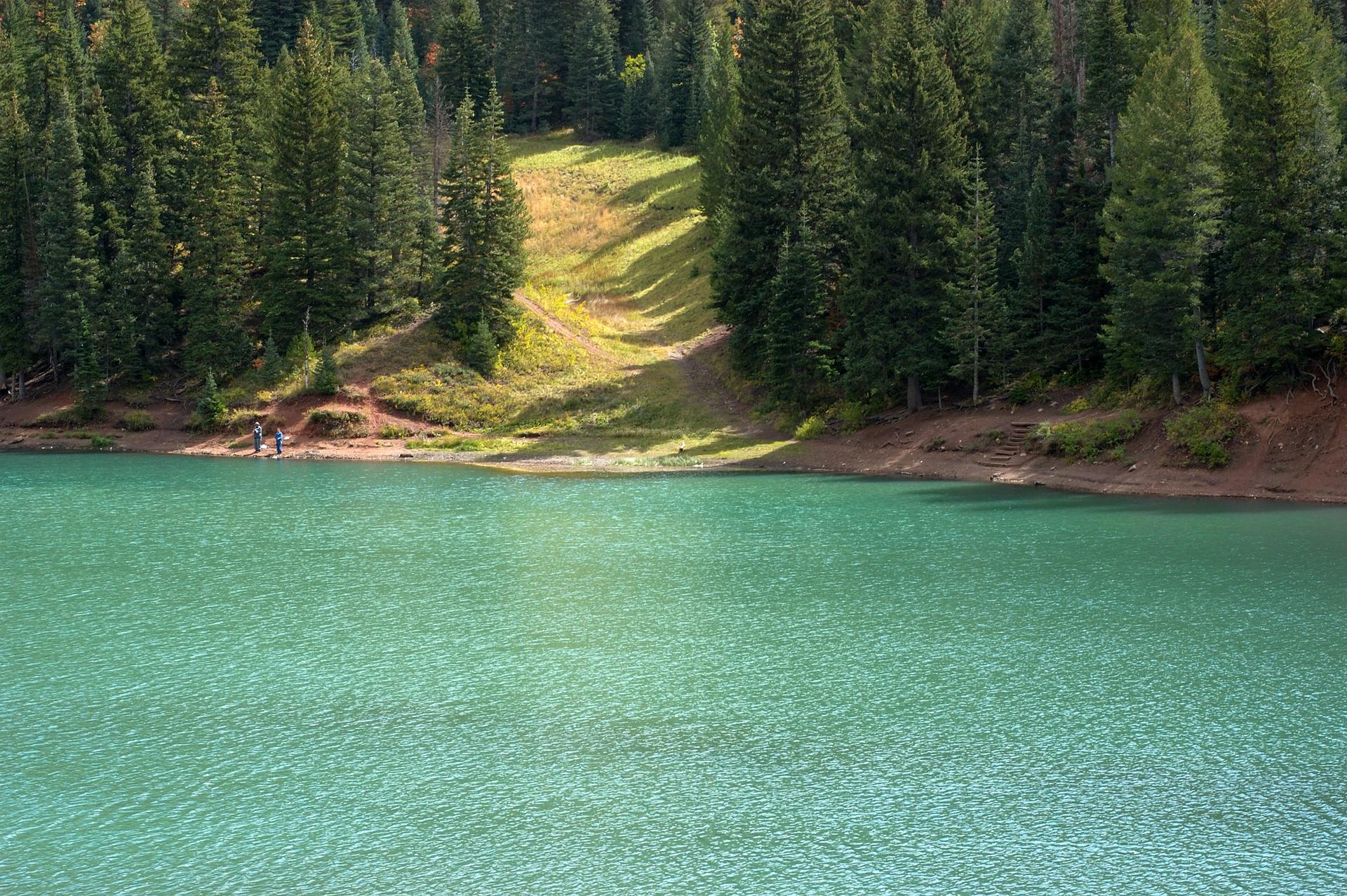 Turquoise lake with dense forest and sunlit hillside, two people by the shore.