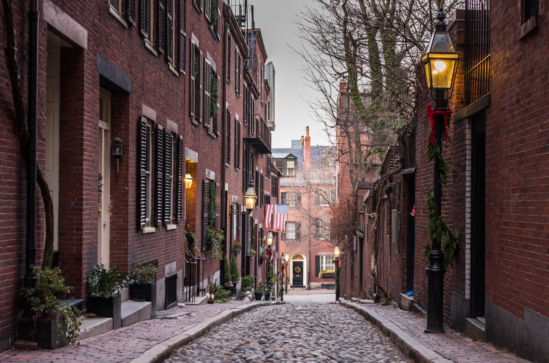 Cobblestone street lined with brick buildings and illuminated gas lamps, overcast.