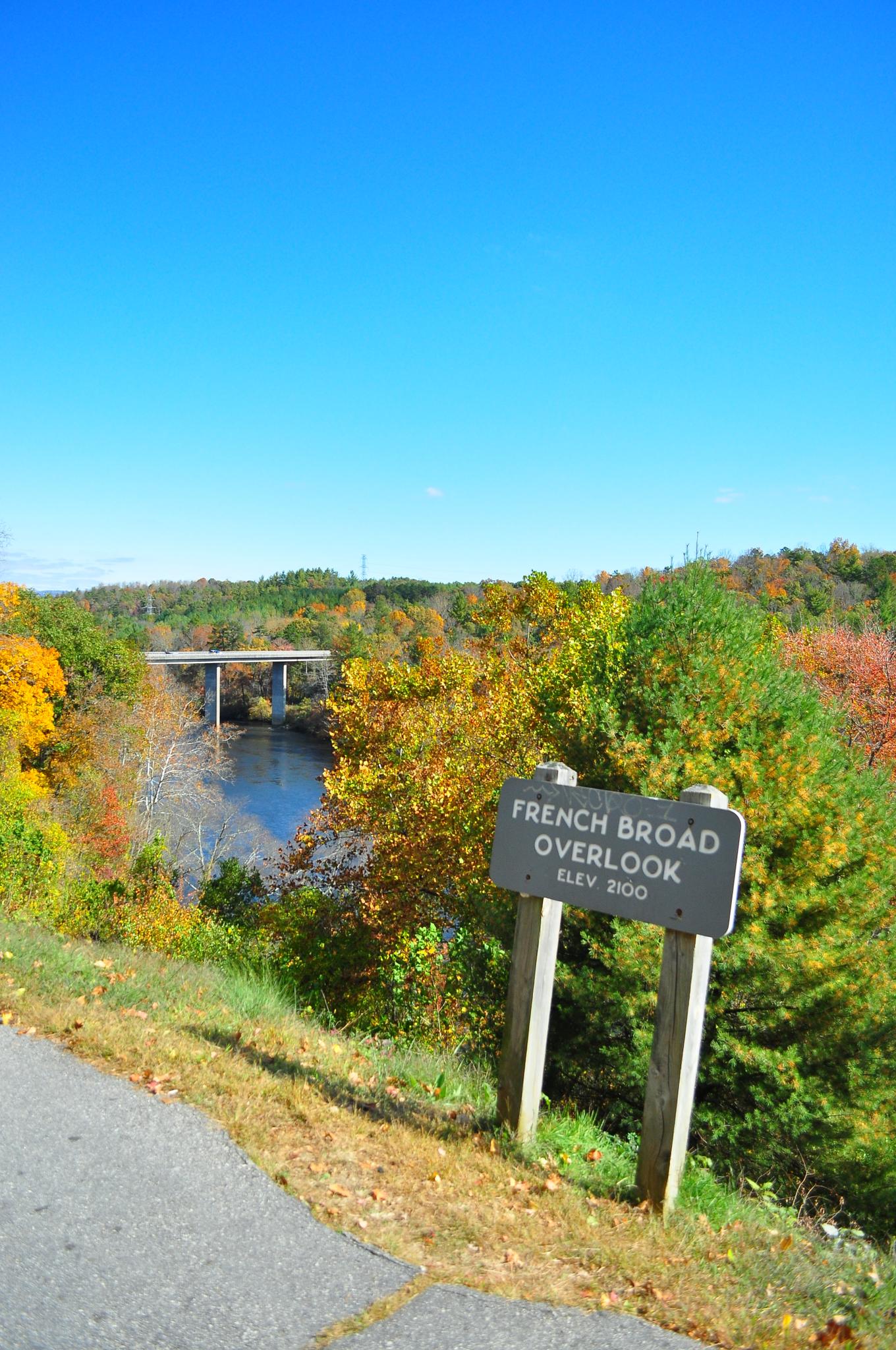 An overlook sign stands above a river, distant bridge, and autumn trees.