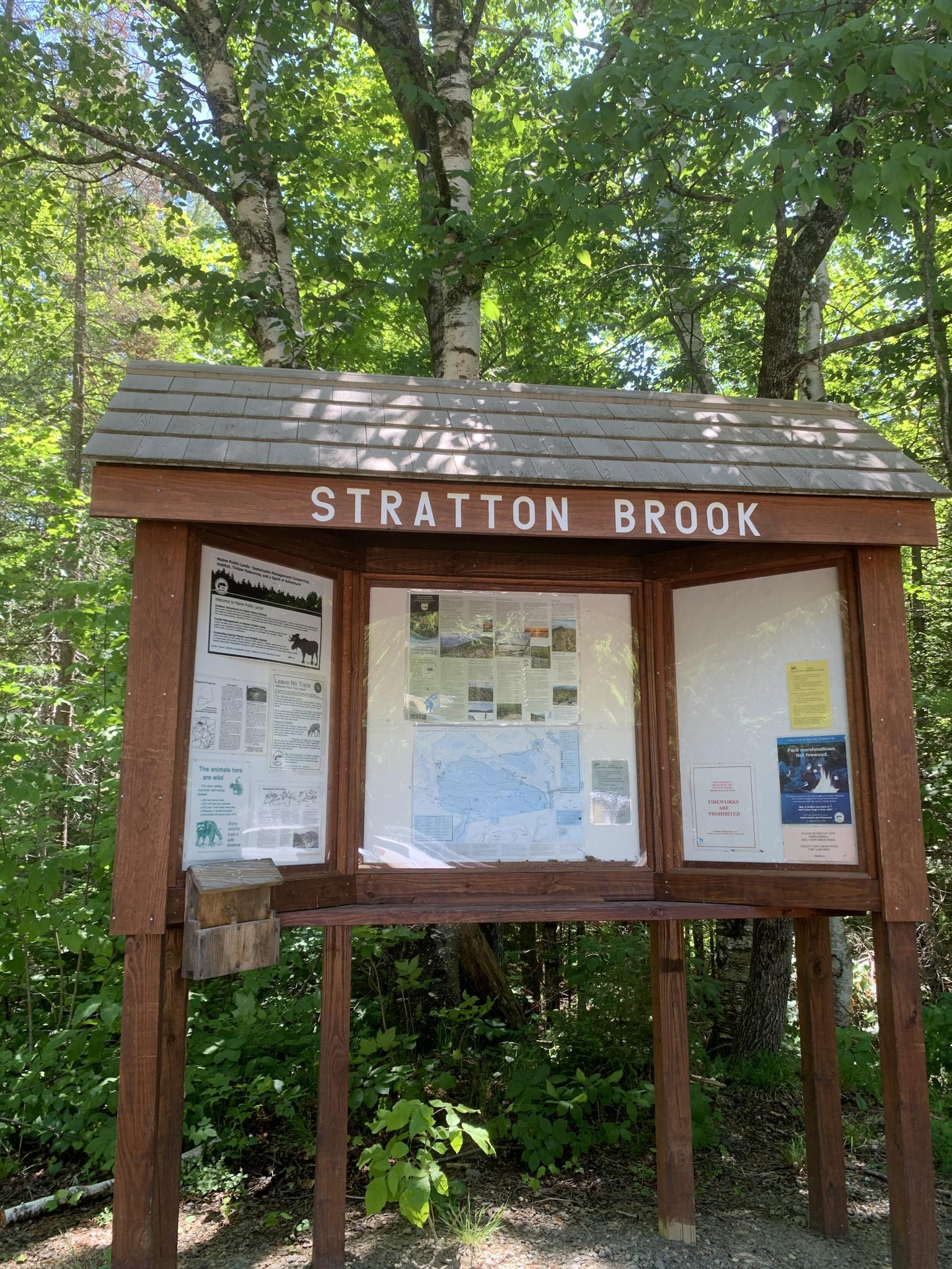 Outdoor information kiosk displaying maps and notices, surrounded by green trees in daylight.
