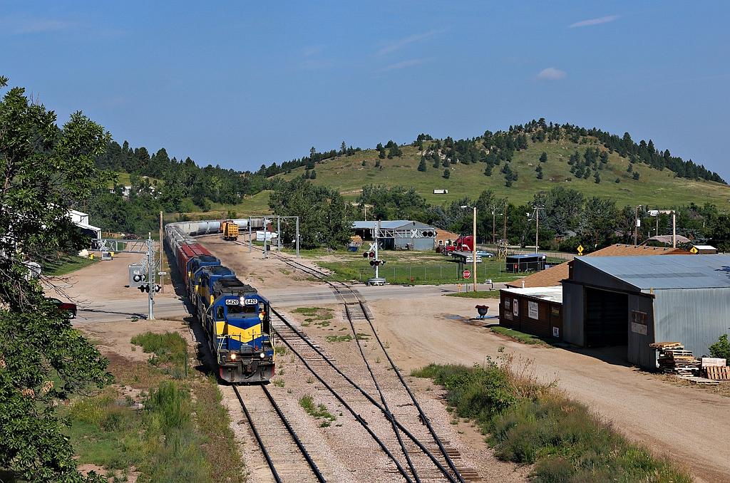 Blue and yellow freight train on tracks, with hills and buildings under bright sun.