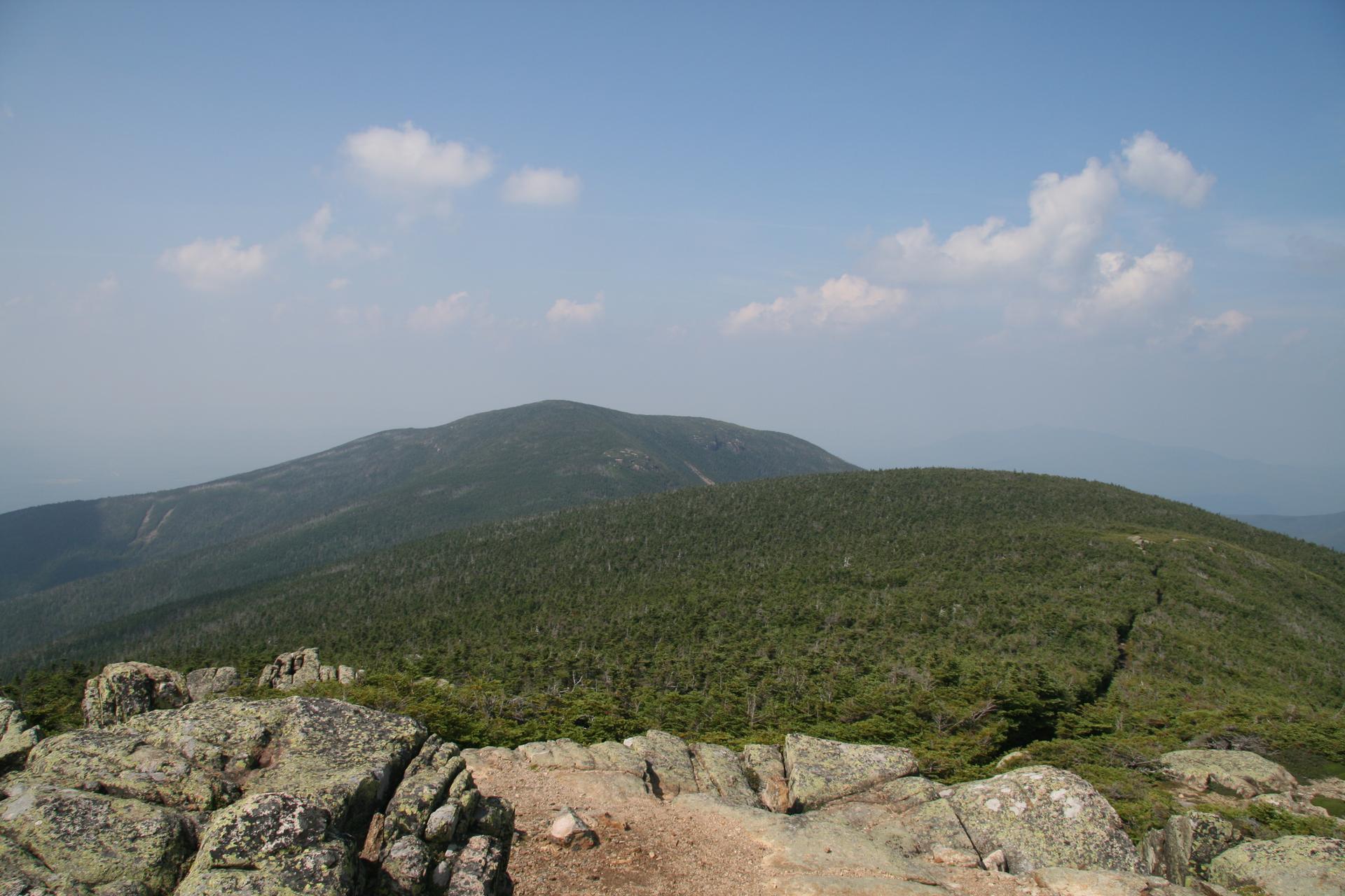Rocky foreground overlooks forested mountain peaks under a bright, cloudy sky.