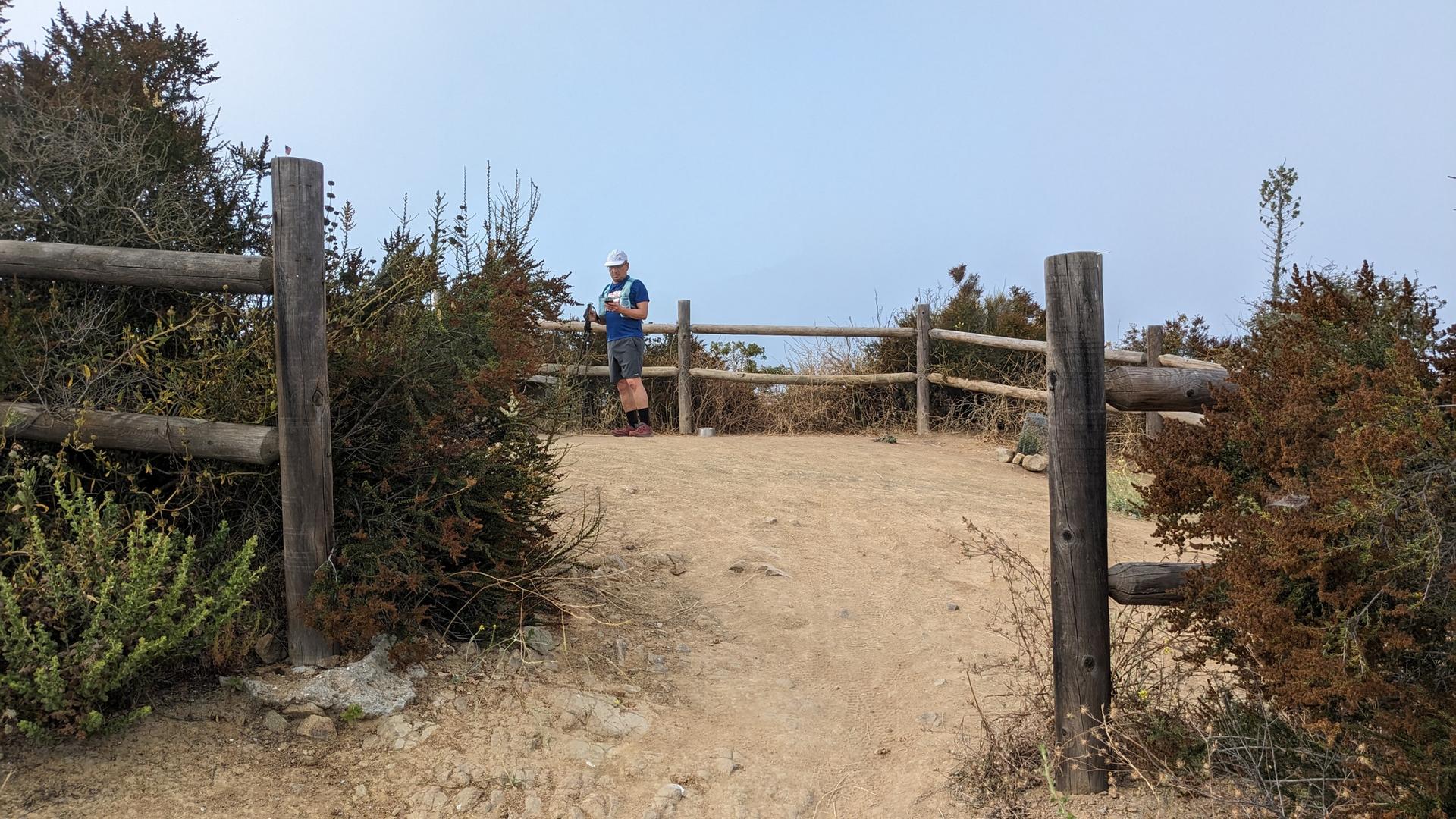 Man standing by a wooden fence on a hilltop, clear sky.