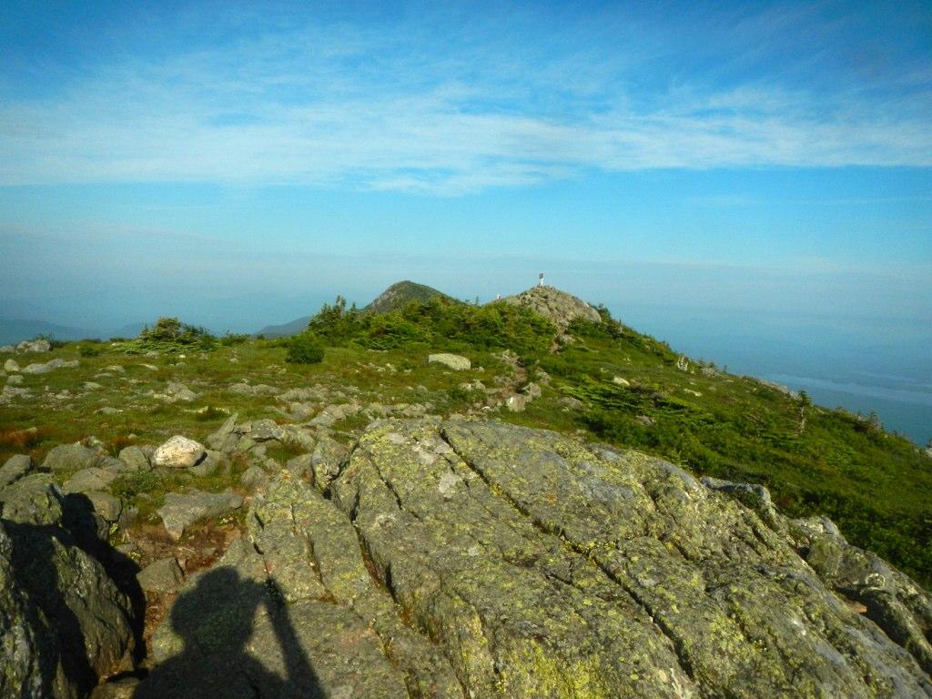 Rocky mountain summit with green slopes, distant peaks, and a person's shadow.