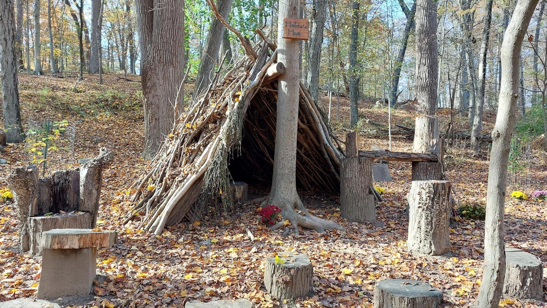 A rustic stick hut surrounded by tree stump seats in an autumn forest.