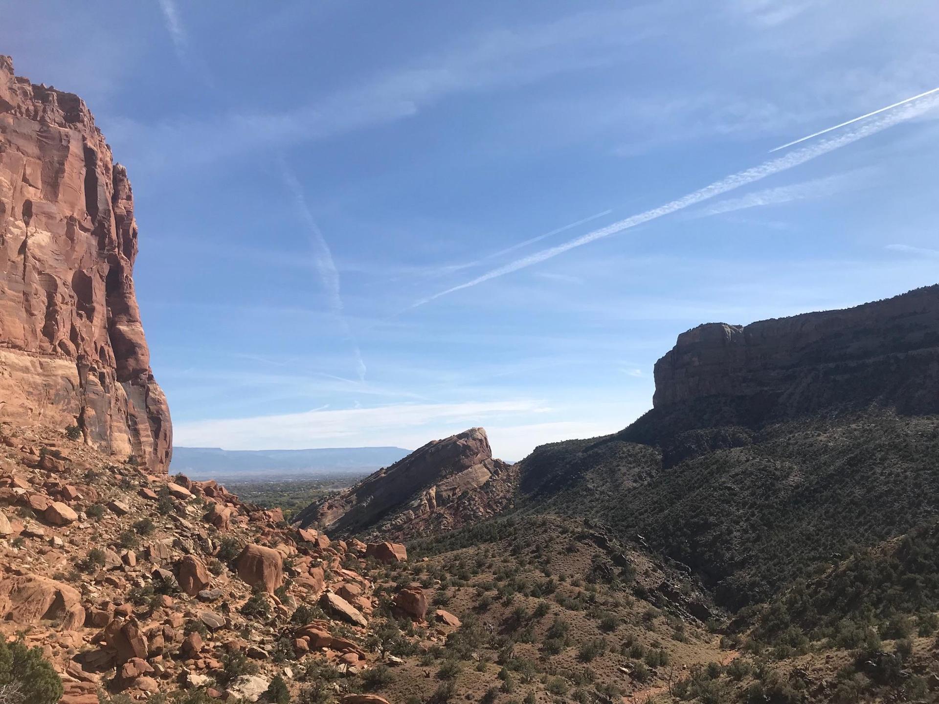 Sunny desert canyon with towering red rock cliffs and distant mountains under blue sky.