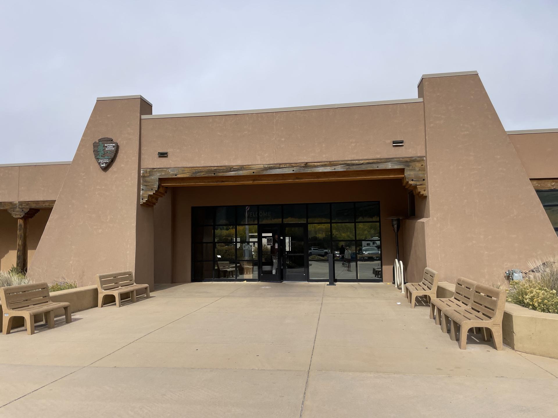 Modern building entrance with large glass doors and outdoor benches under an overcast sky.