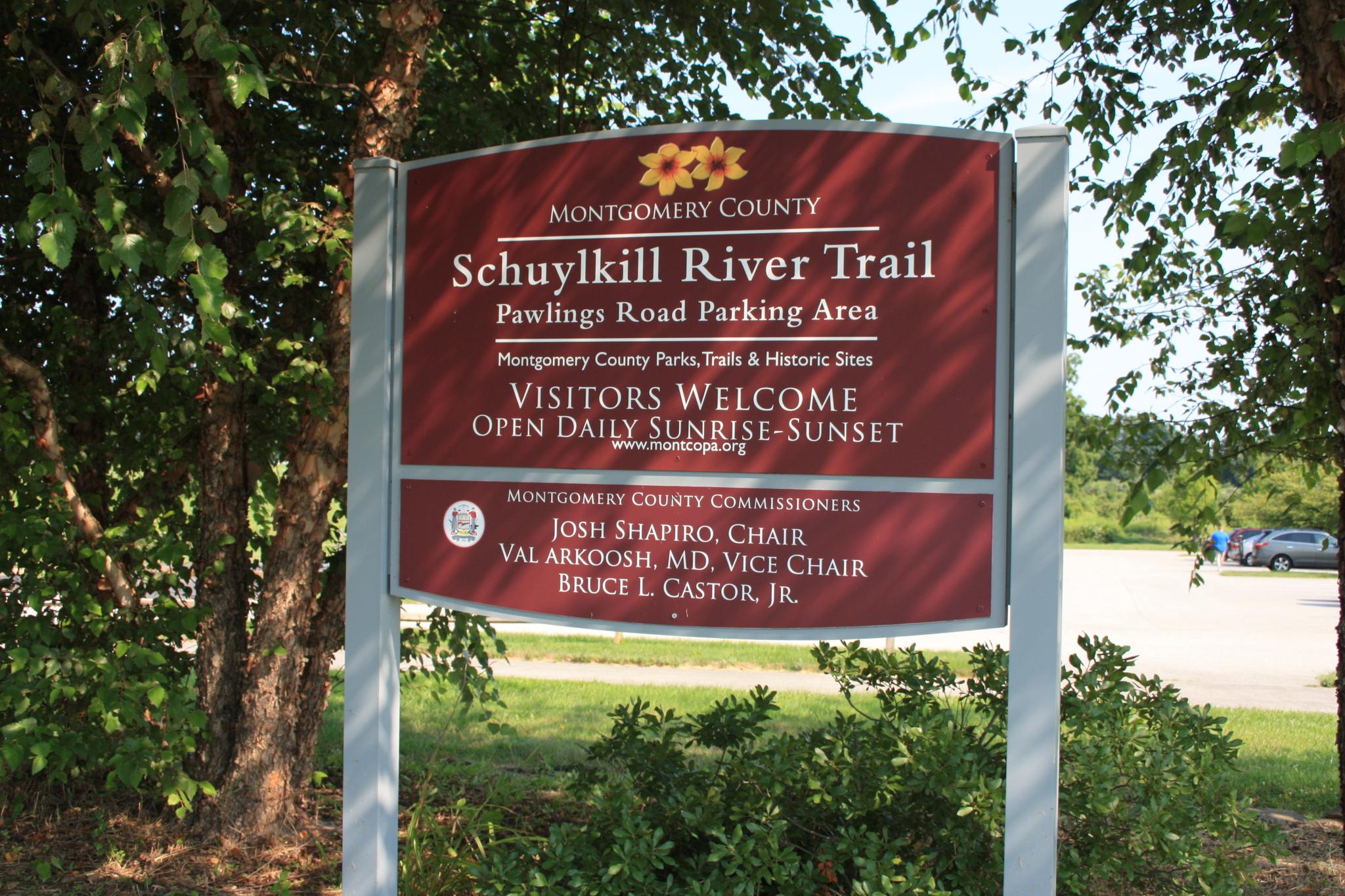 A large red sign stands among green trees, with a parking lot visible in daylight.
