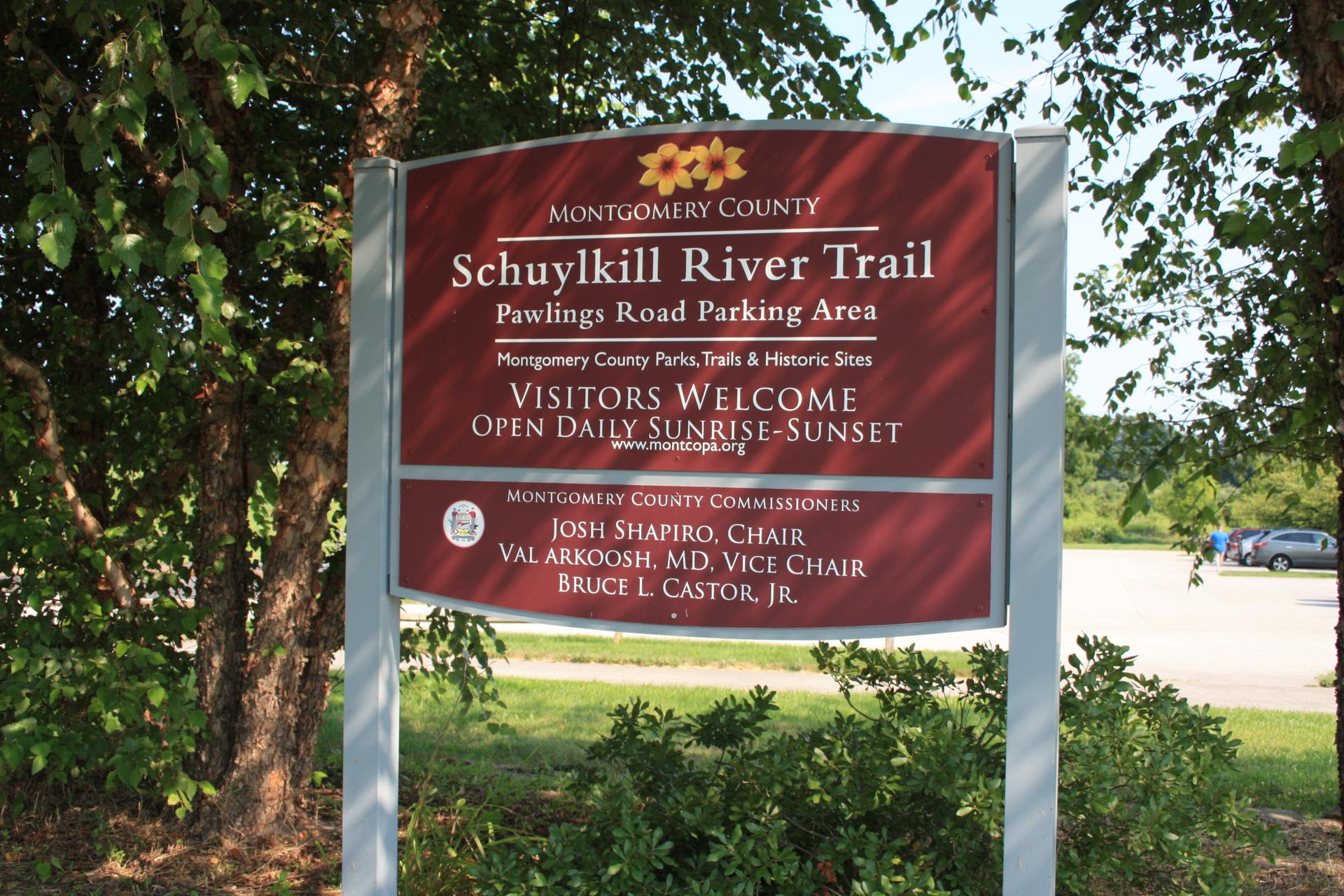 A large red sign stands among green trees, with a parking lot visible in daylight.